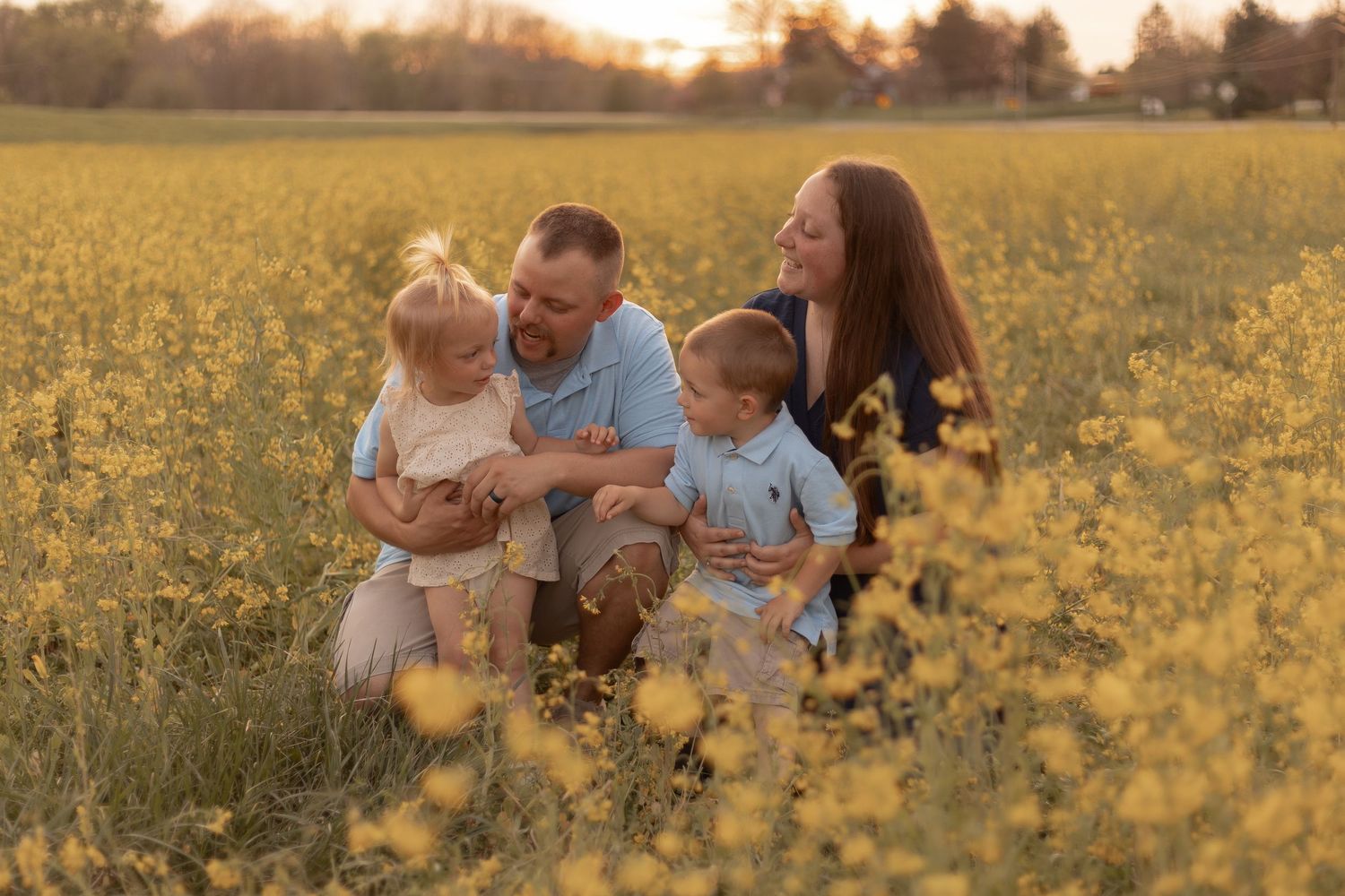Family of four smiling together in a golden yellow wildflower field at sunset.