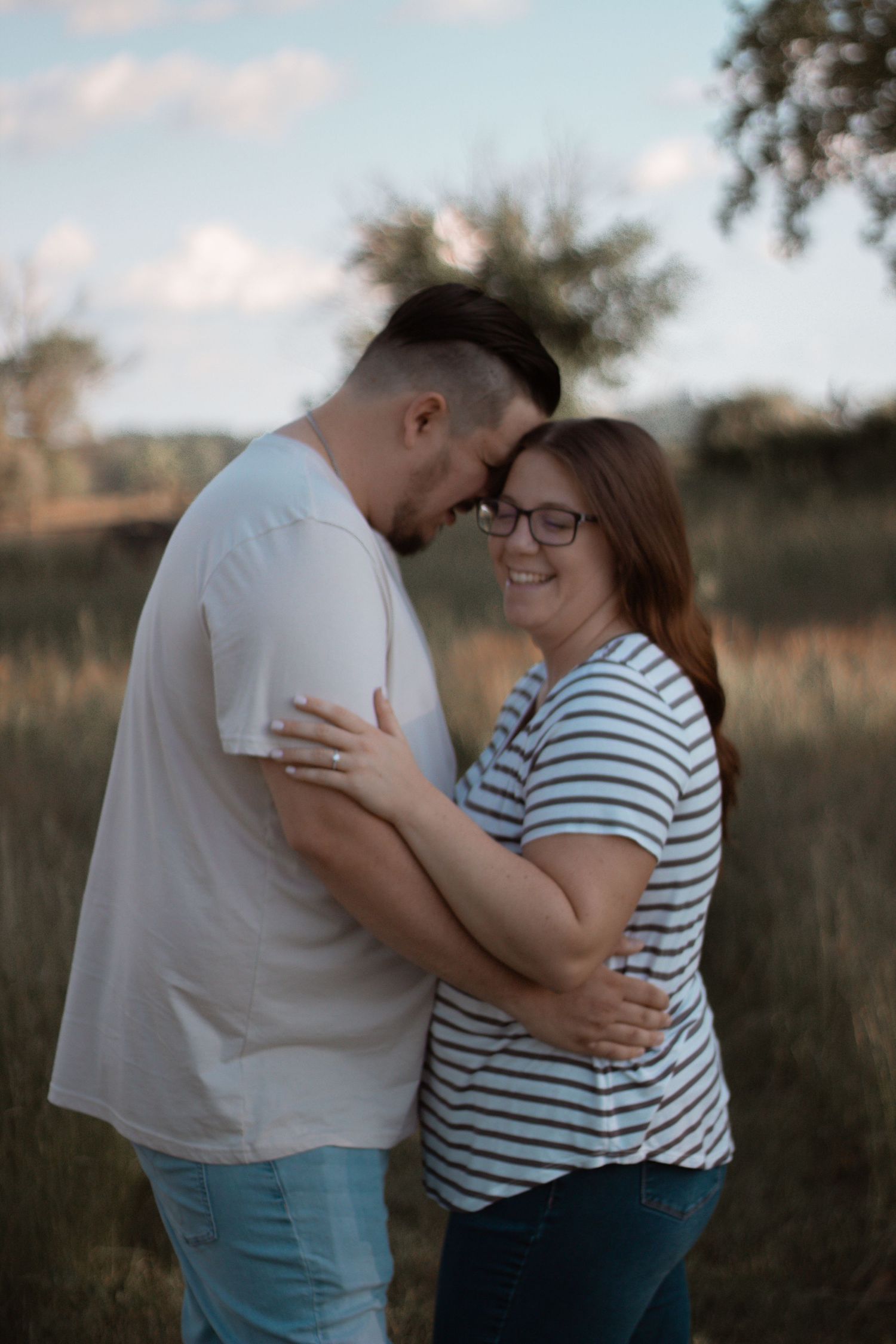 A couple shares an intimate embrace in a field during golden hour, wearing casual striped and white clothing.