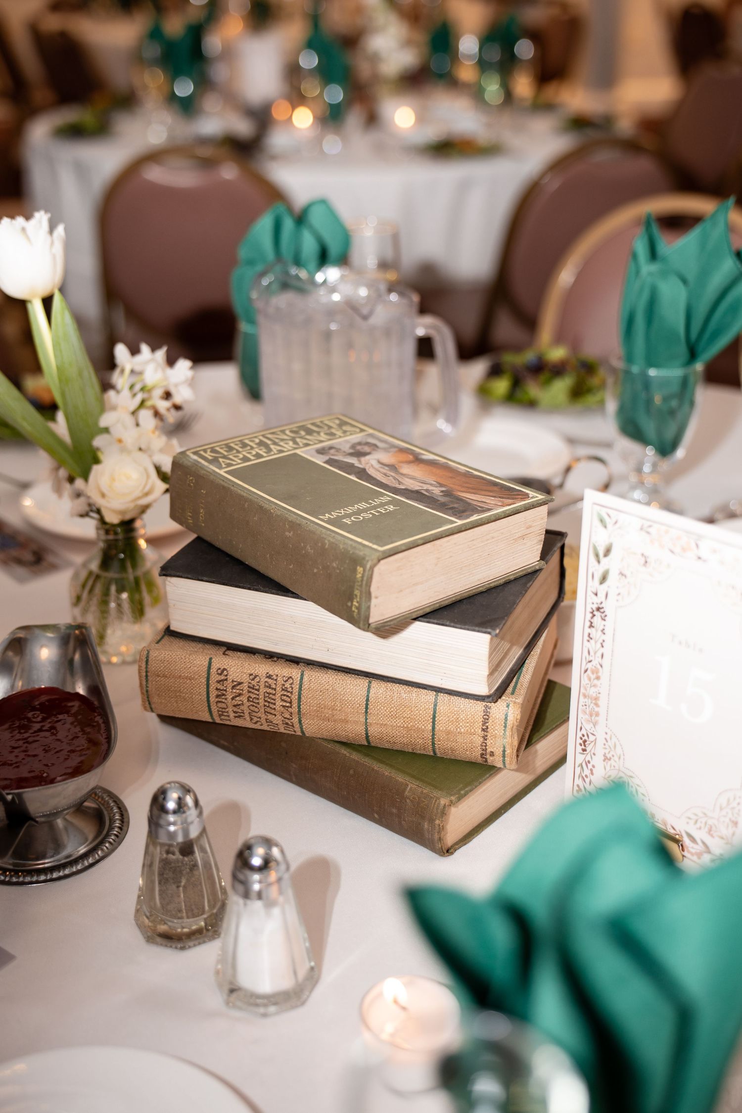 Stack of vintage books used as elegant wedding reception centerpiece with teal napkins and floral accents.