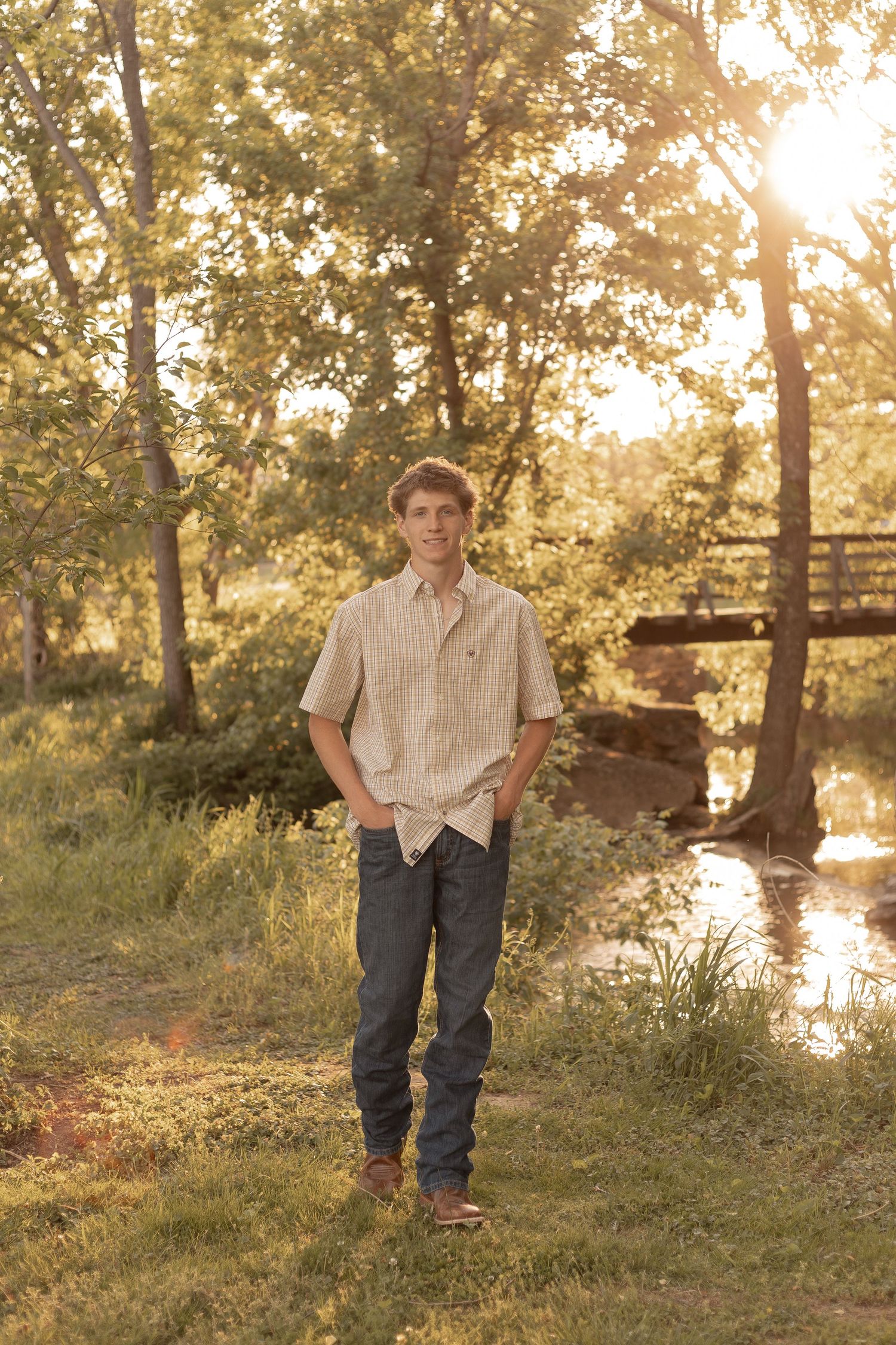 Young man in plaid shirt and jeans stands in sunlit park near creek and wooden bridge.