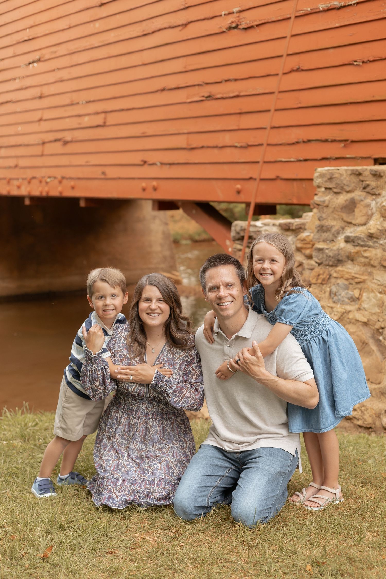 Family of four posing happily outdoors near a red covered bridge, smiling for a portrait on green grass.