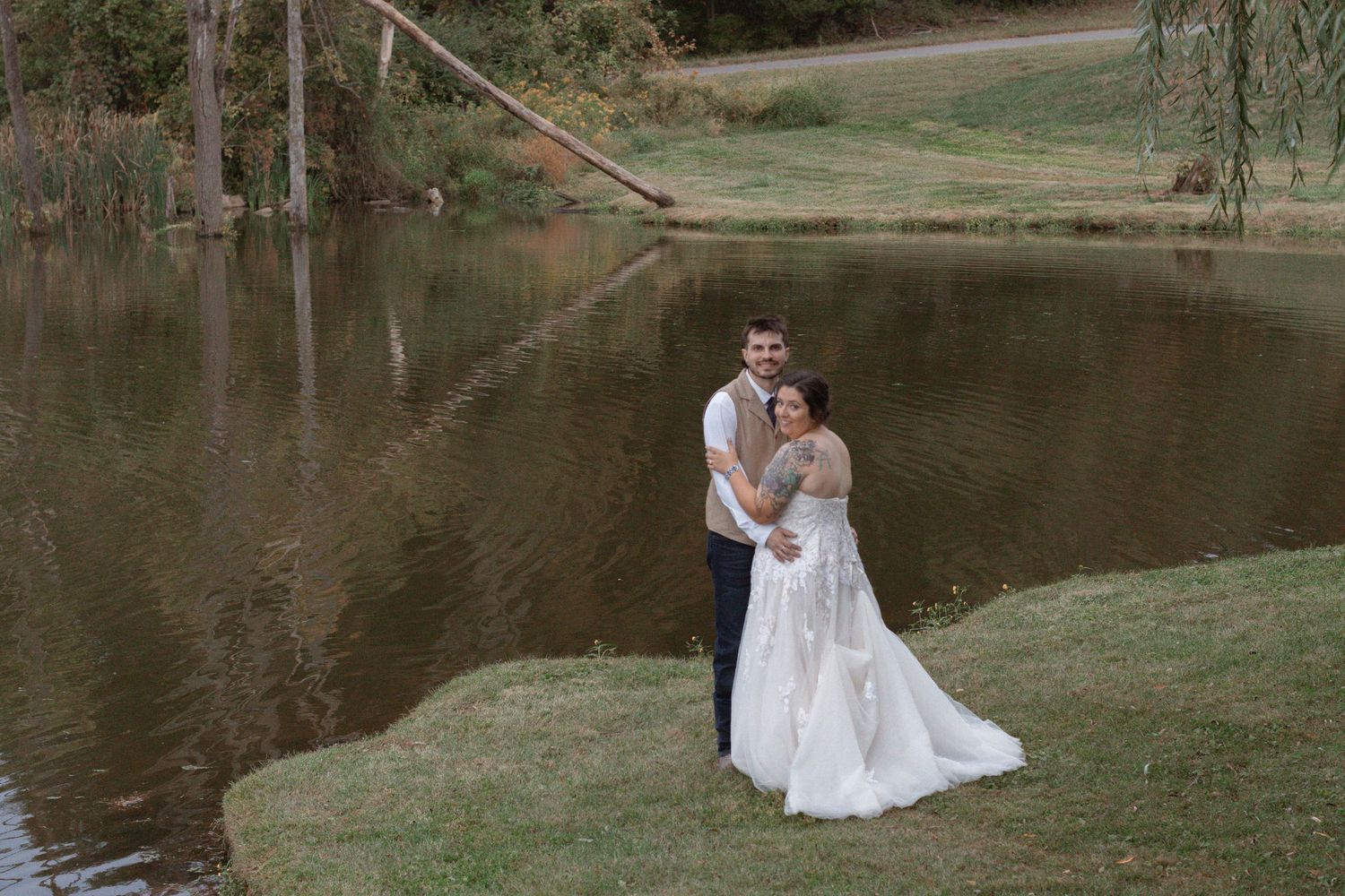 A couple in wedding attire shares intimate moments by a serene lake surrounded by lush greenery and trees.