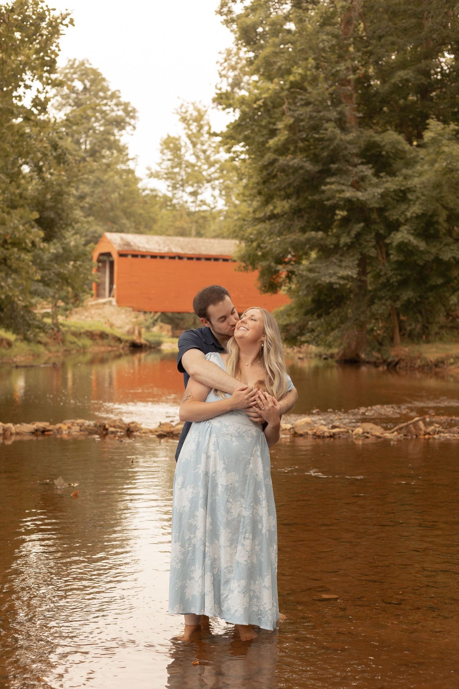Couple embracing romantically in a shallow creek with a red covered bridge and lush green trees in the background.