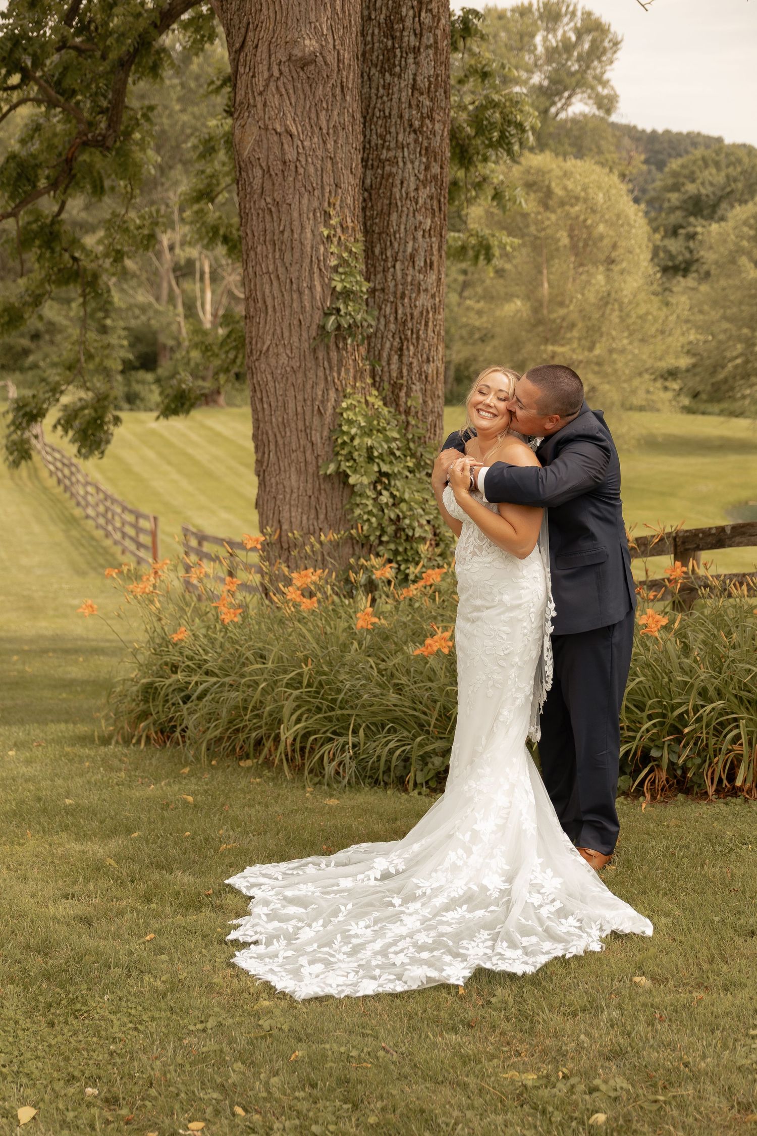 Bride in lace gown and groom in navy suit share a romantic kiss outdoors near a large tree with orange flowers.