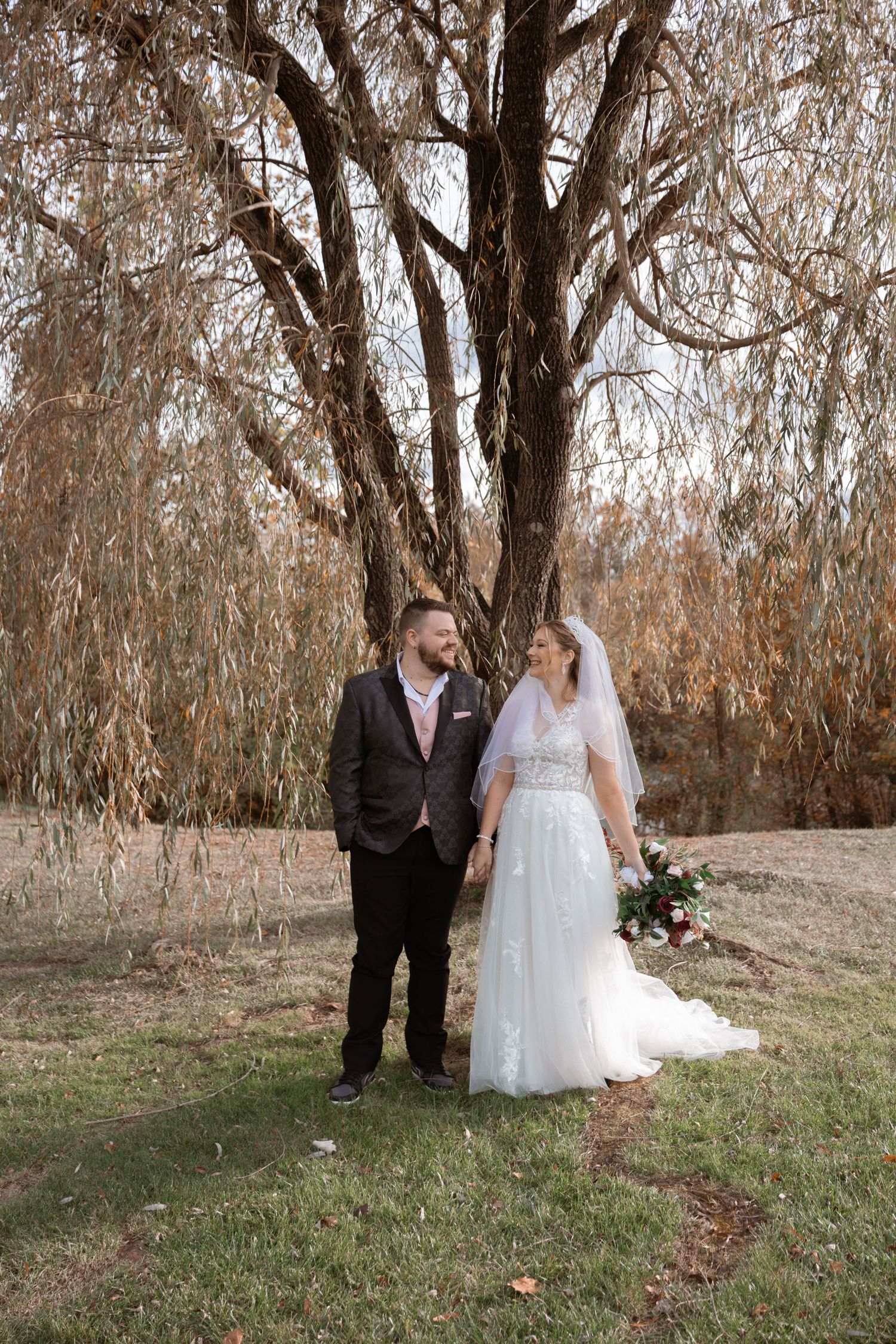 A romantic wedding couple stands among autumn trees with fallen leaves in a natural outdoor setting.