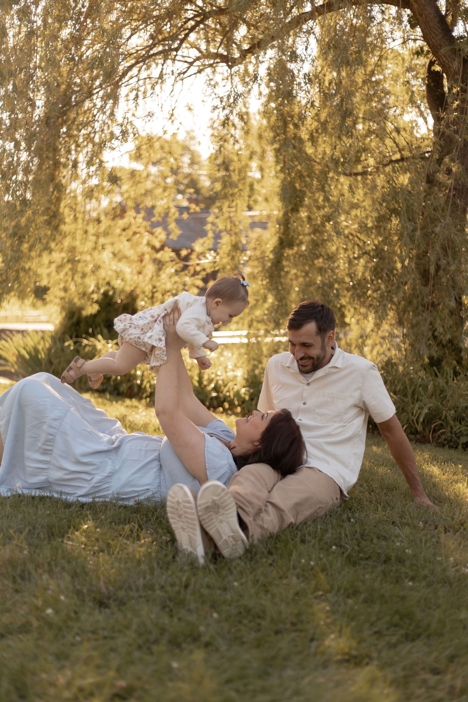 A happy family relaxing on grass under a willow tree, with mother lifting baby while father watches lovingly.