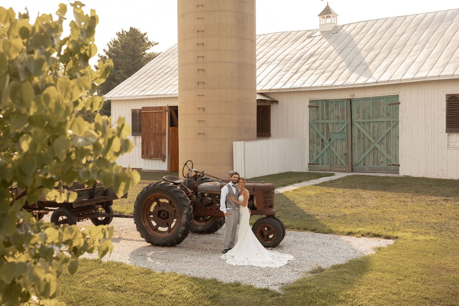 Bride and groom pose beside a vintage tractor in front of a rustic white barn with green doors.