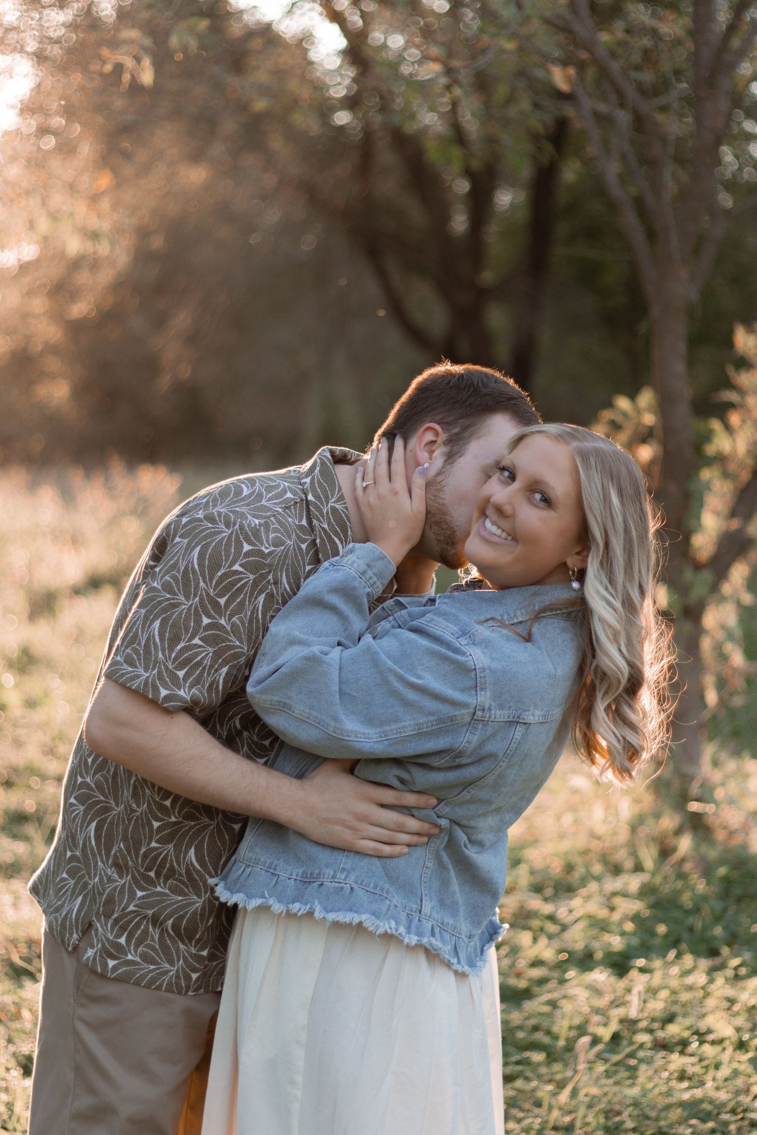 A couple embraces tenderly in a sun-dappled outdoor setting with golden light filtering through trees.
