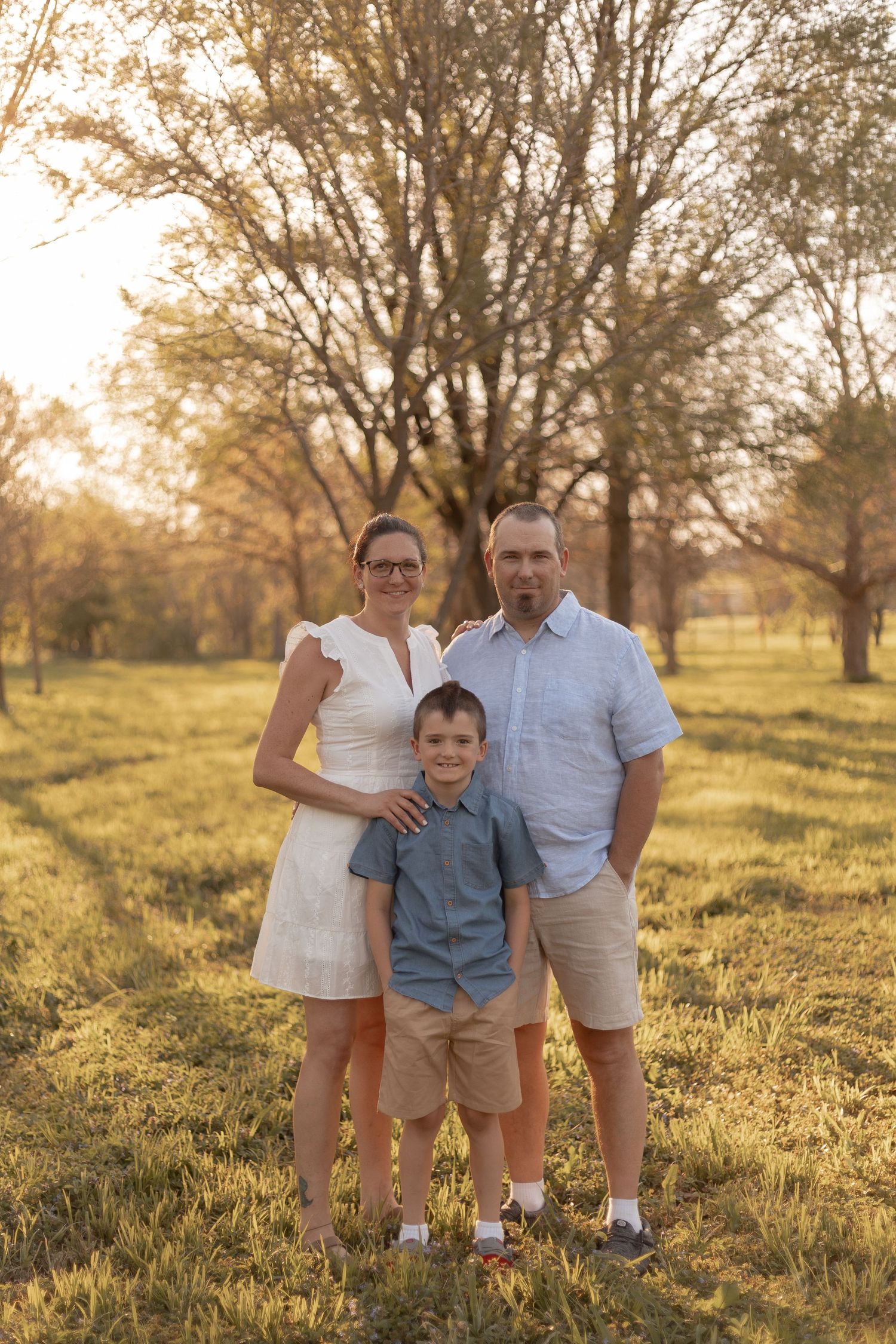 Family of three posing outdoors at golden hour, parents and young boy smiling in a sunlit park with trees.