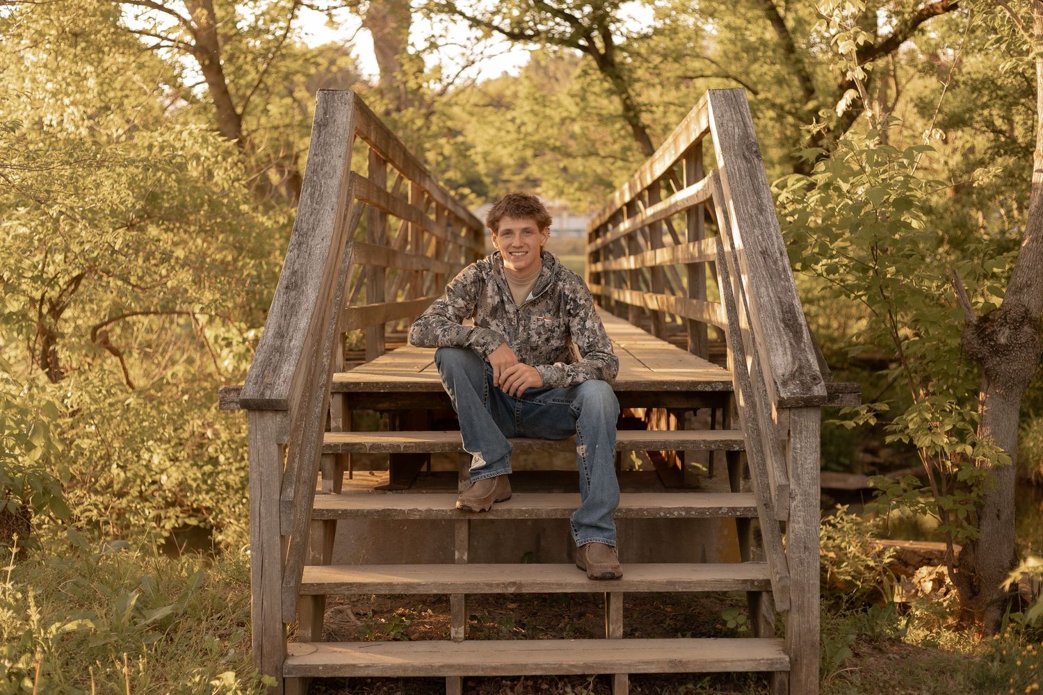 Young man in camo shirt and jeans sitting on wooden bridge steps surrounded by lush green trees.