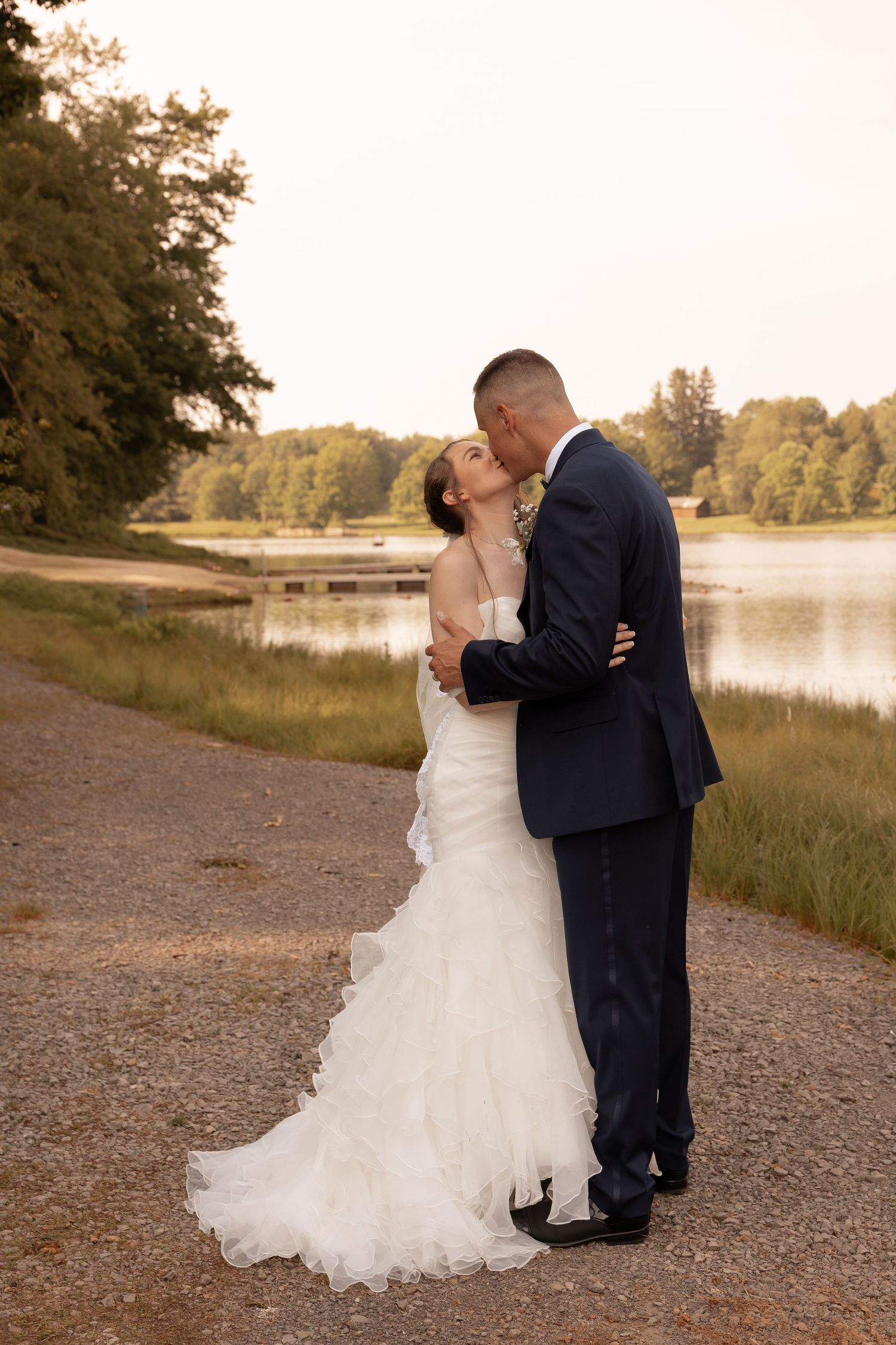 Bride and groom share a romantic kiss by a serene lake at sunset, she in a white gown, he in a navy suit.