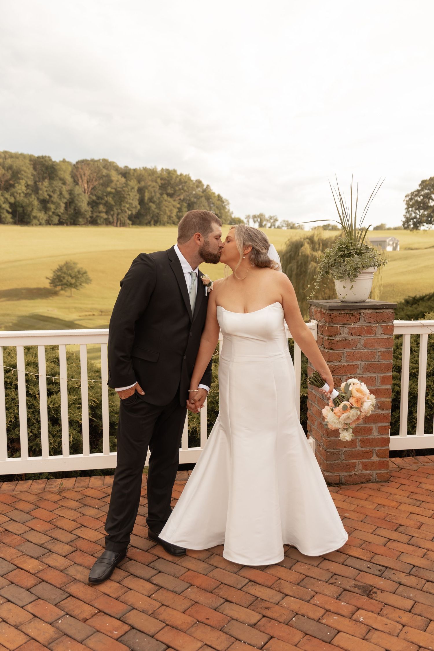 Bride and groom share a kiss on a brick terrace, she holds a bouquet in a white gown, he wears a black suit.