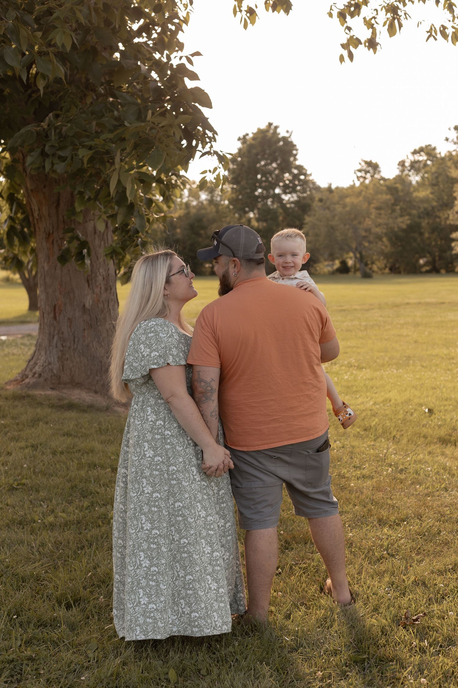 A young family with a toddler enjoys a sunny day in a green park, mom in floral dress and dad in orange shirt.