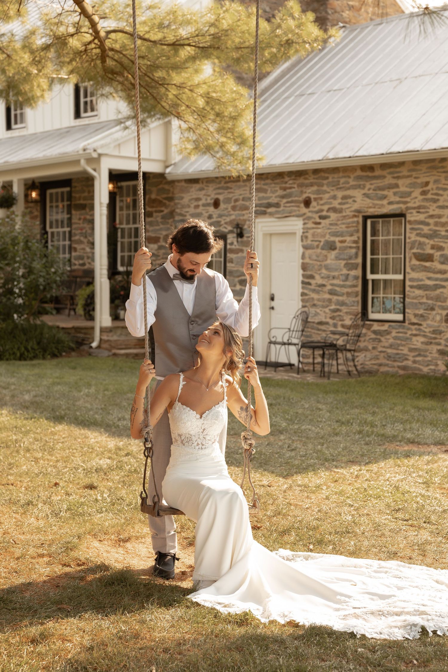 Bride sitting on a rope swing while groom stands behind her in front of a rustic stone farmhouse on their wedding day.