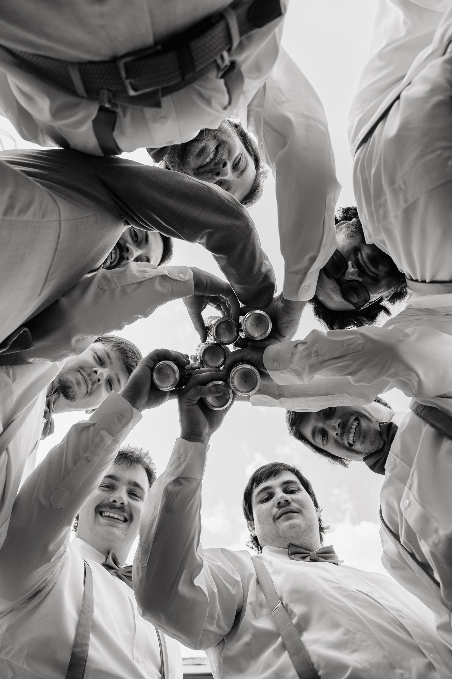 Black and white photo of groomsmen in a huddle toasting with beer cans, shot from below.