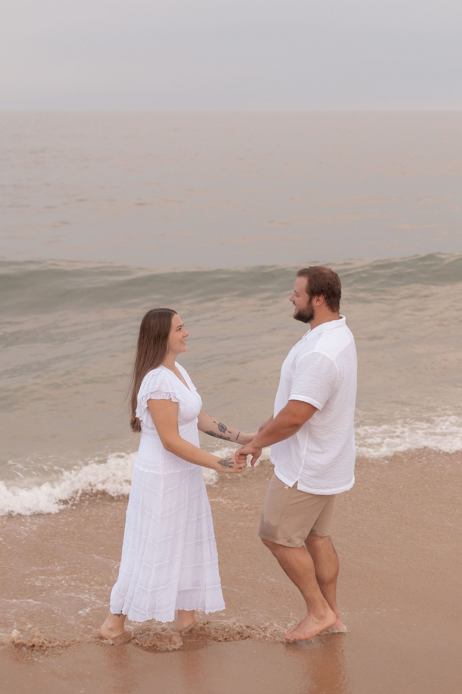 Couple holding hands on a misty beach, woman in white dress and man in white shirt and khaki shorts, waves behind them.