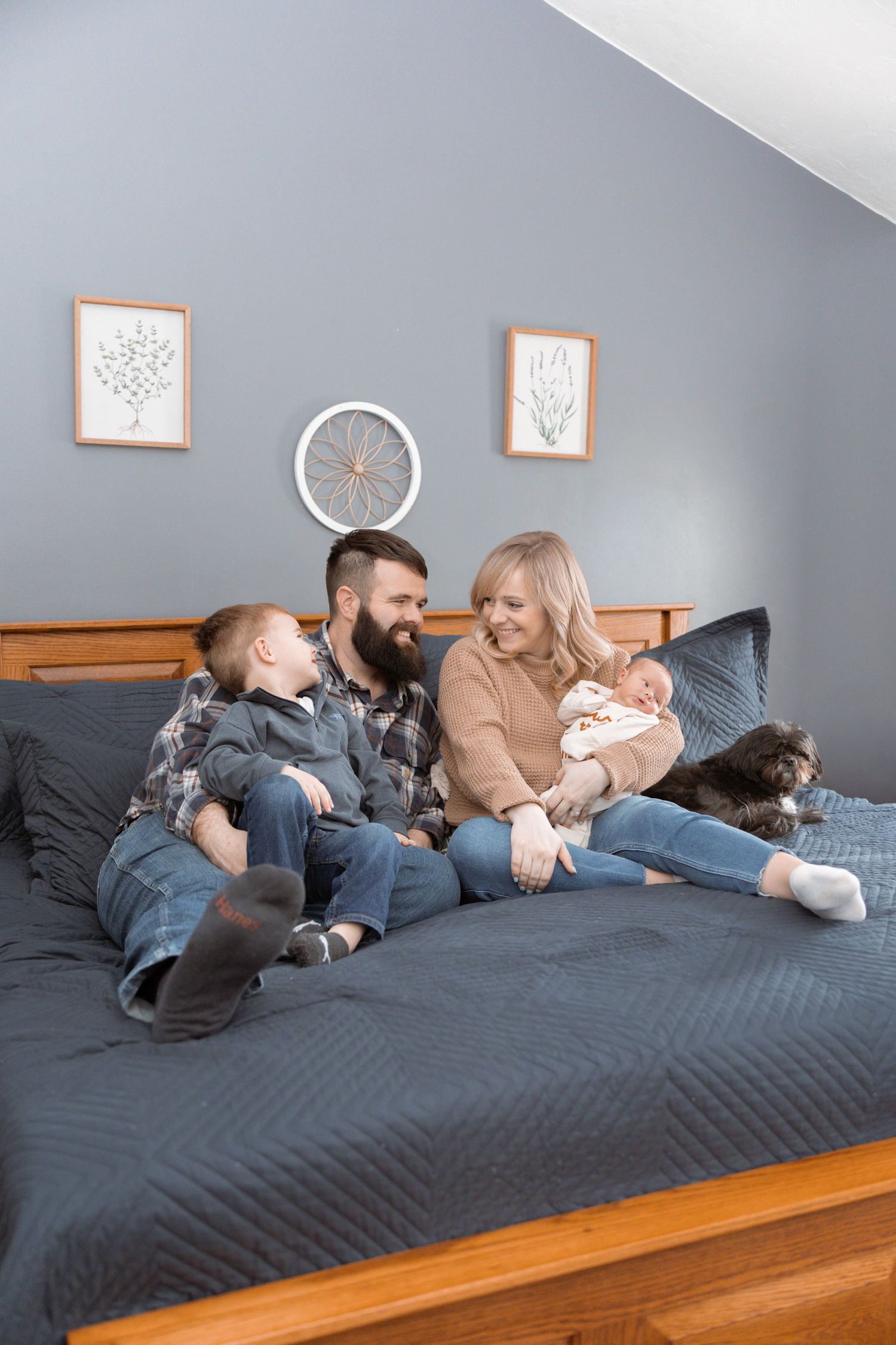 A happy family with a bearded dad, blonde mom, toddler, newborn baby, and dog relaxing together on a blue bed.