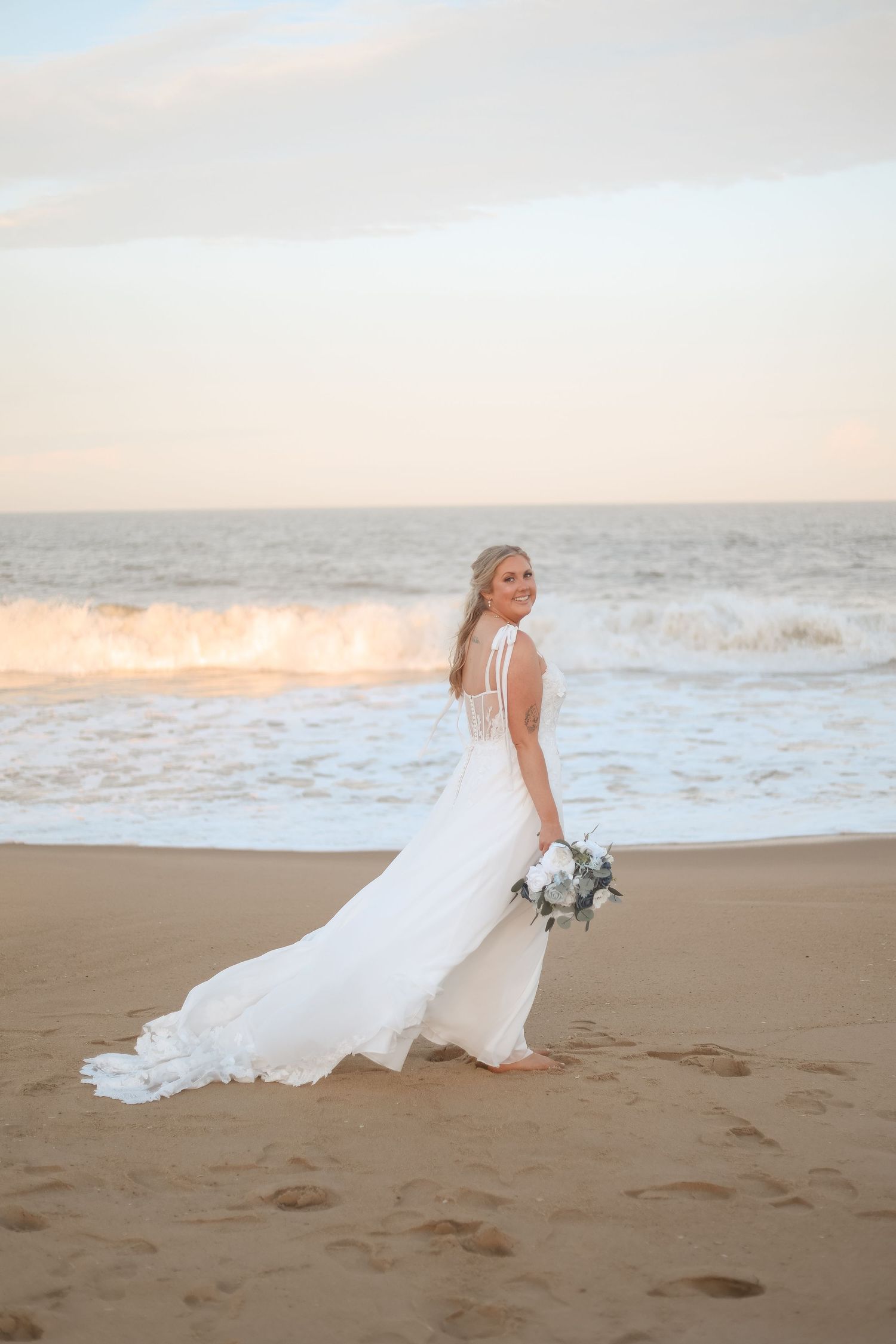 Bride in white gown holding bouquet on sandy beach at sunset with ocean waves behind her.