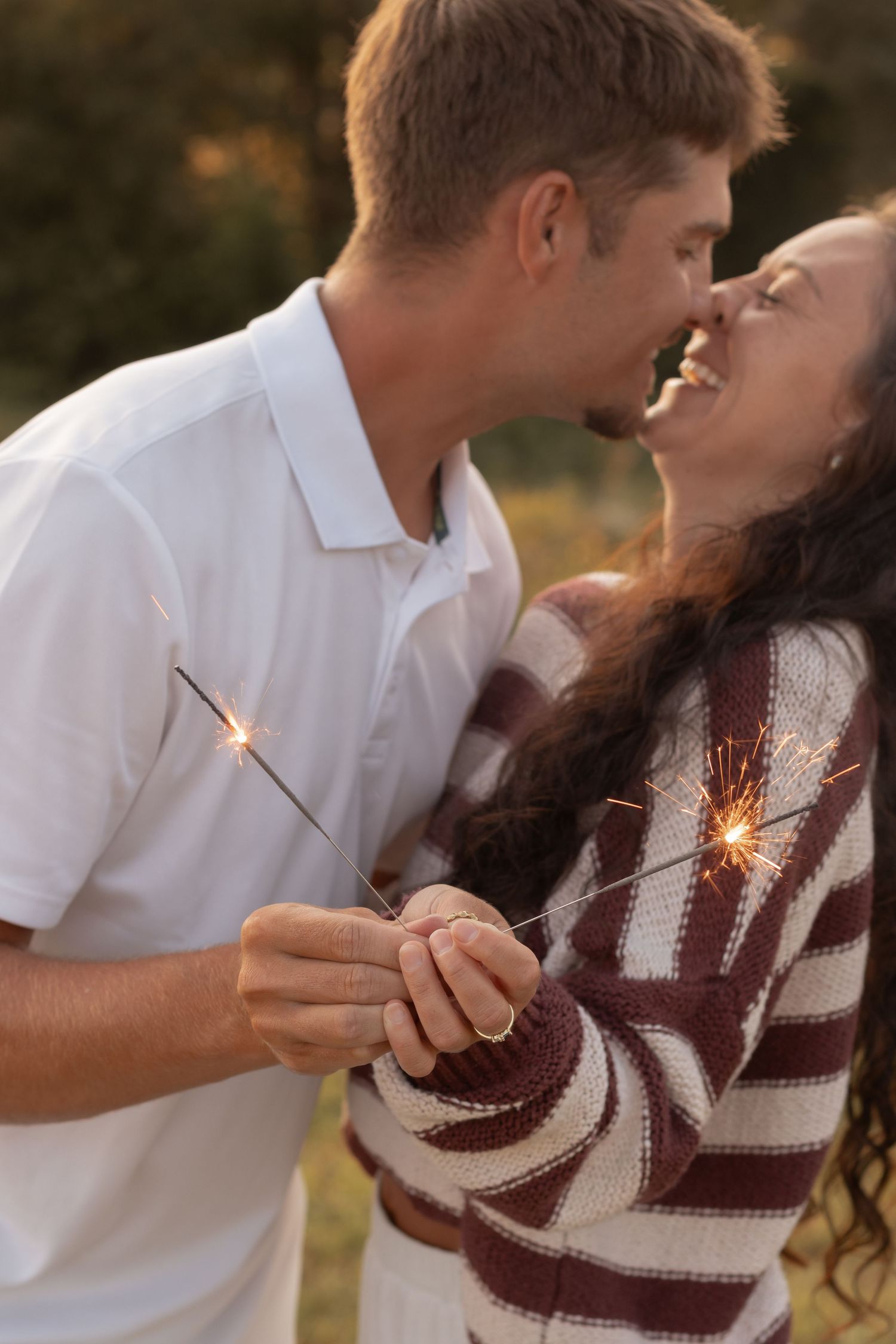 A joyful couple sharing a kiss while holding sparklers outdoors, wearing casual sweater and white polo shirt.