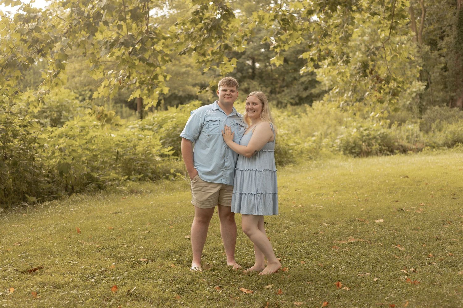 Couple in matching blue outfits posing barefoot on green grass with lush trees in background.