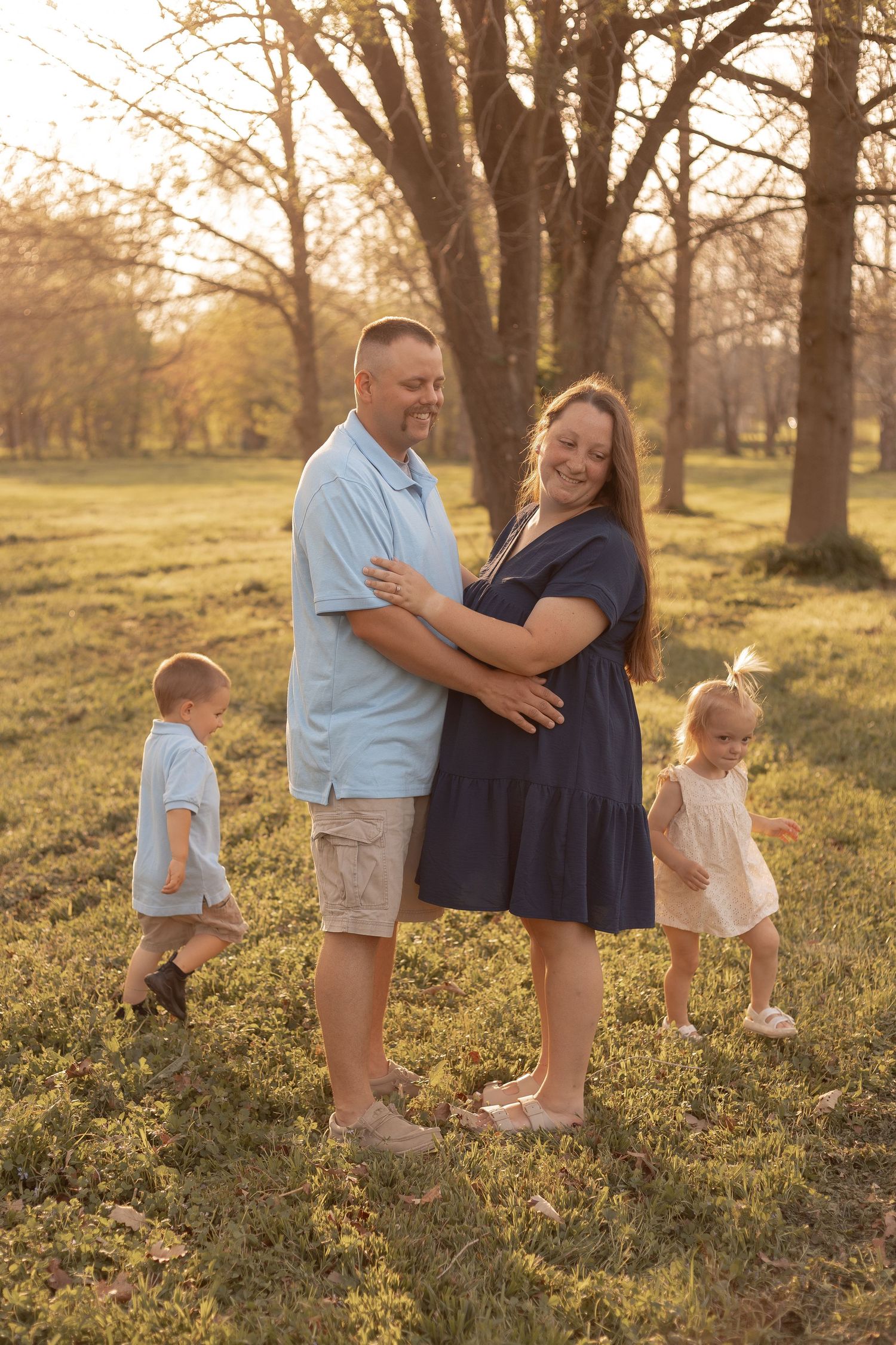 A pregnant woman and man embrace outdoors at golden hour while two young children play around them in a park.