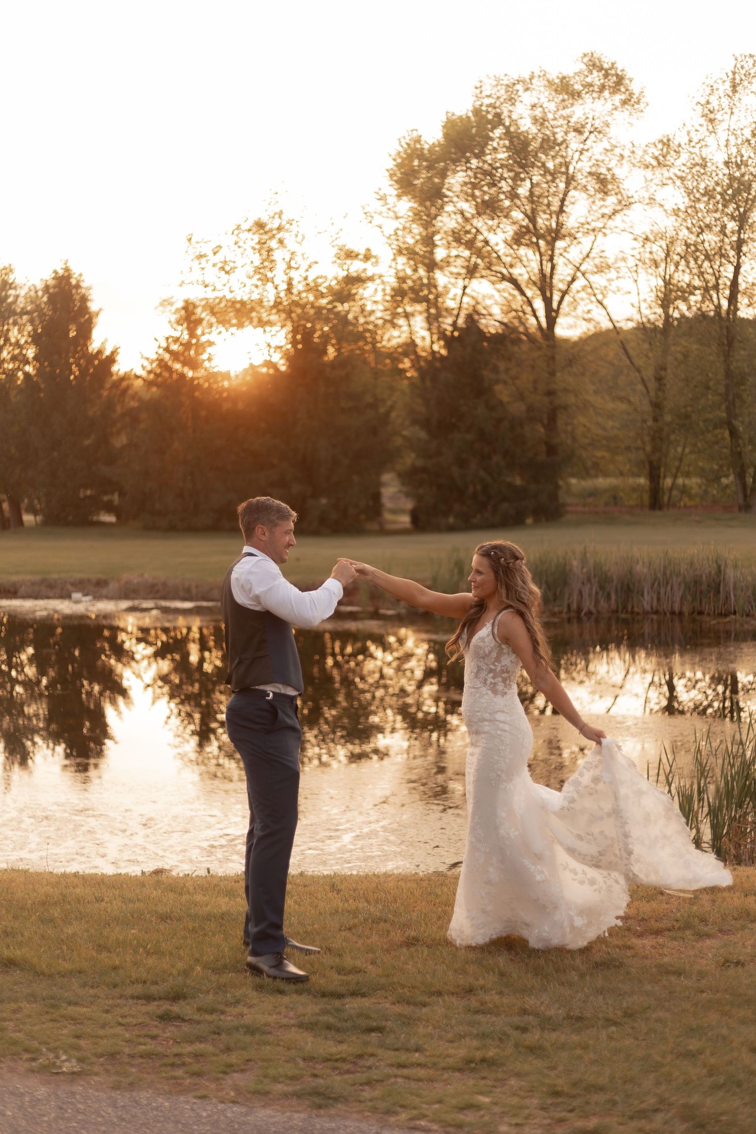 Bride and groom dancing by a sunlit pond at golden hour, surrounded by lush trees reflecting on the water.