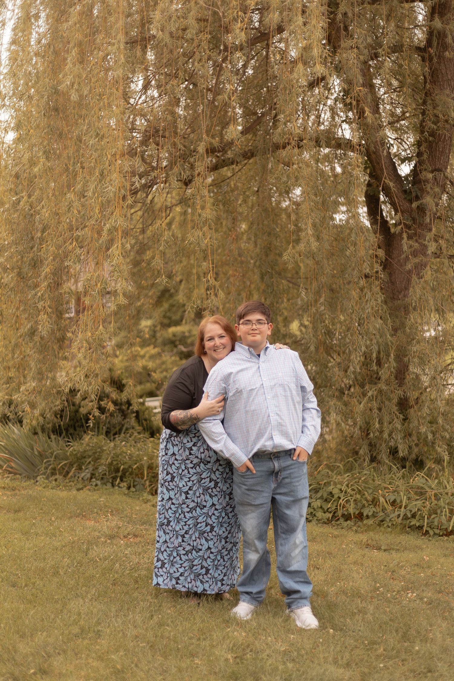 A woman and teenage boy pose together under a weeping willow tree on green grass outdoors.
