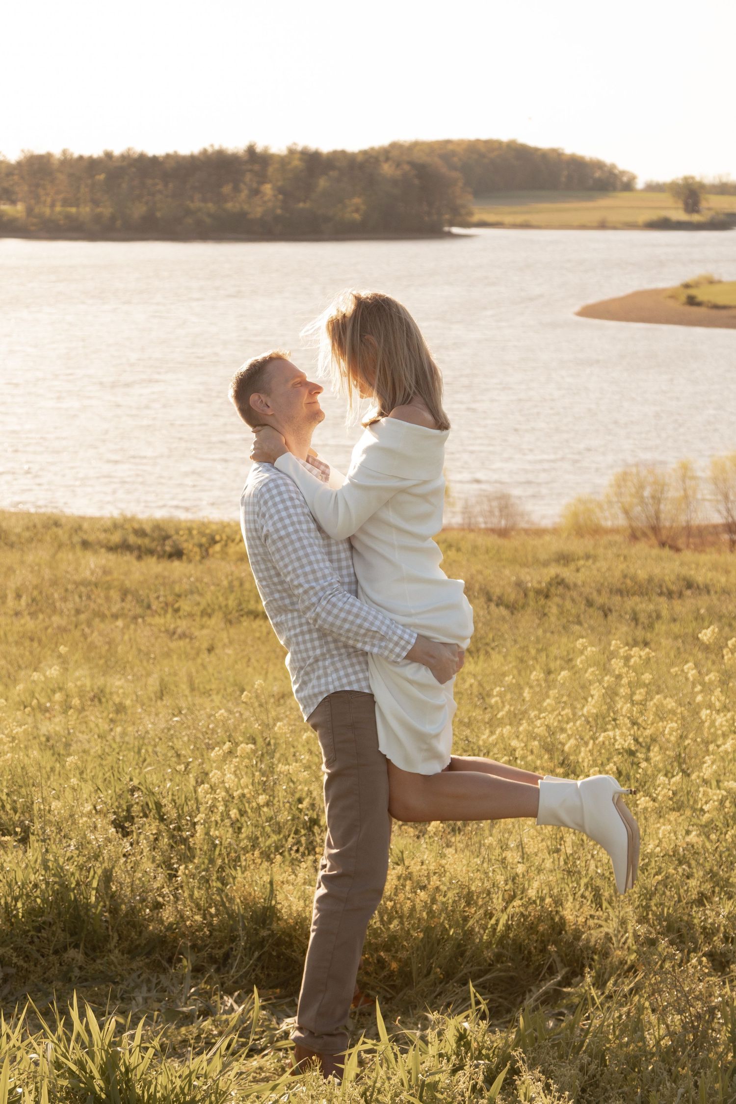 Couple sharing a romantic moment at sunset by a lake, man lifting woman in a golden field.