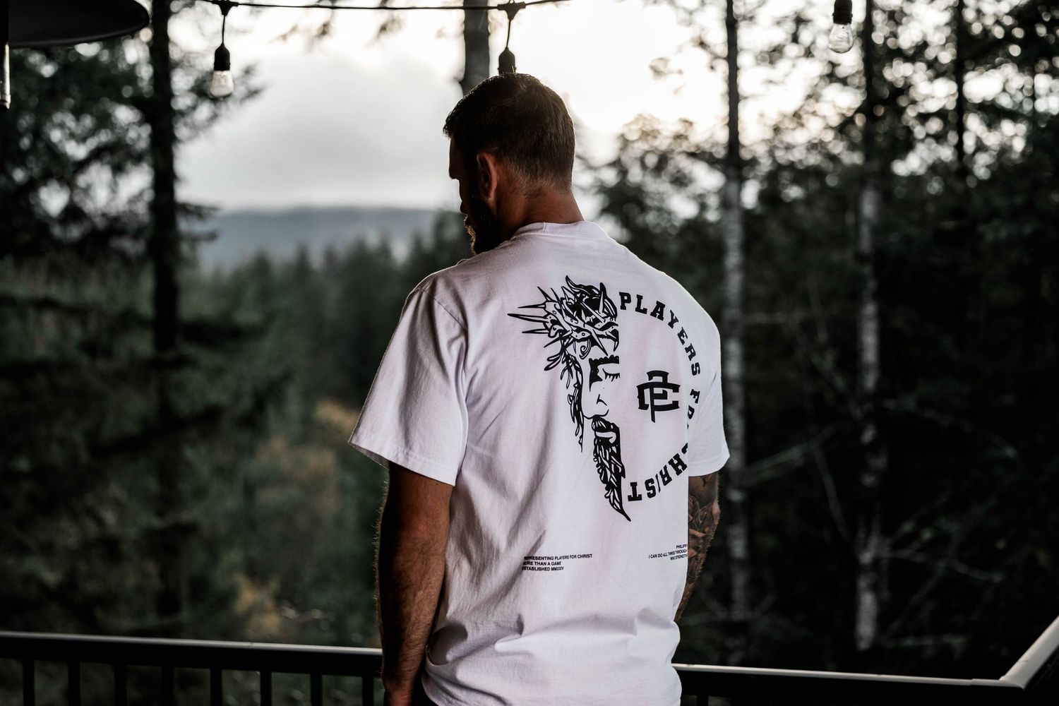 Person wearing white t-shirt with graphic design stands on balcony overlooking forest landscape.