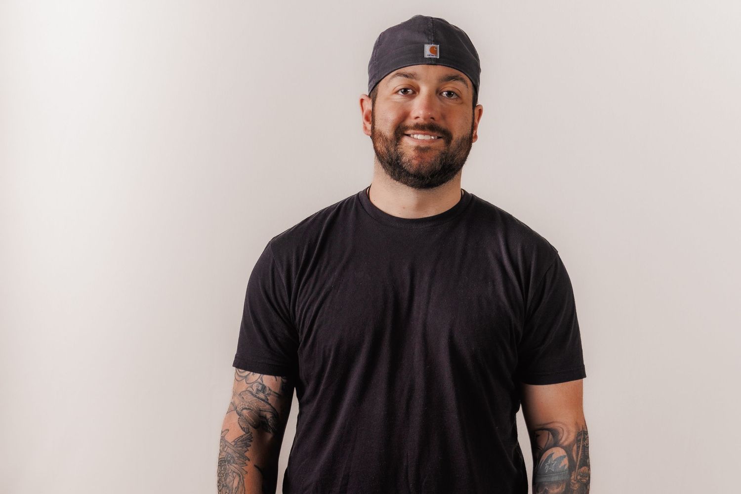Man in black t-shirt and baseball cap smiling warmly against neutral background.