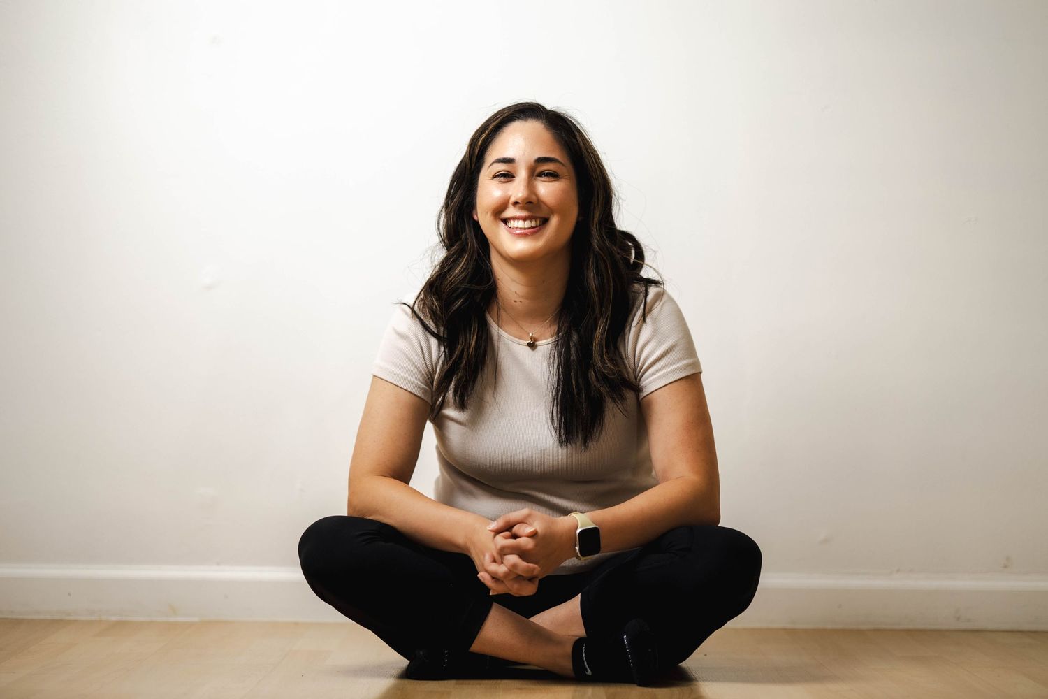 Person sitting cross-legged on wooden floor wearing casual attire against white wall.