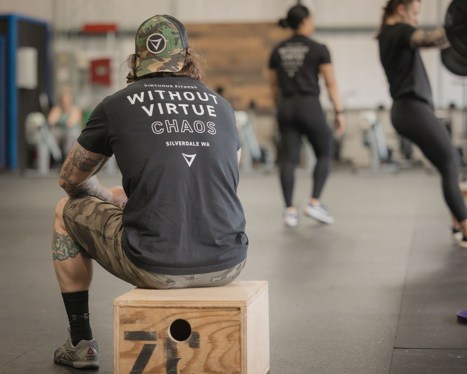 Person in black t-shirt with text sitting on wooden box in gym setting.