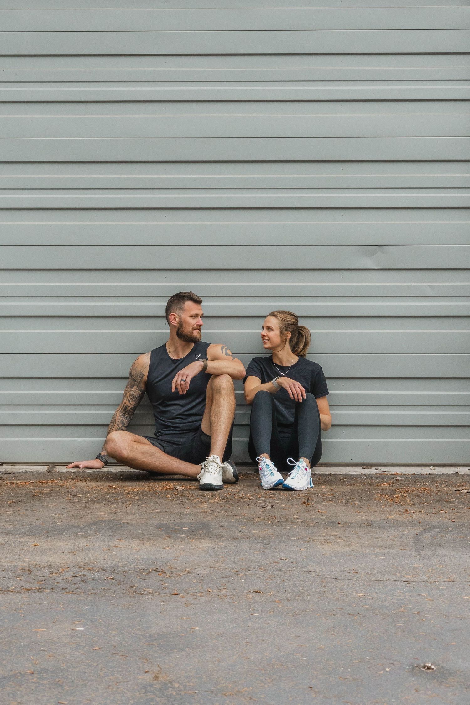 Two people sitting and chatting against a grey metal garage door.