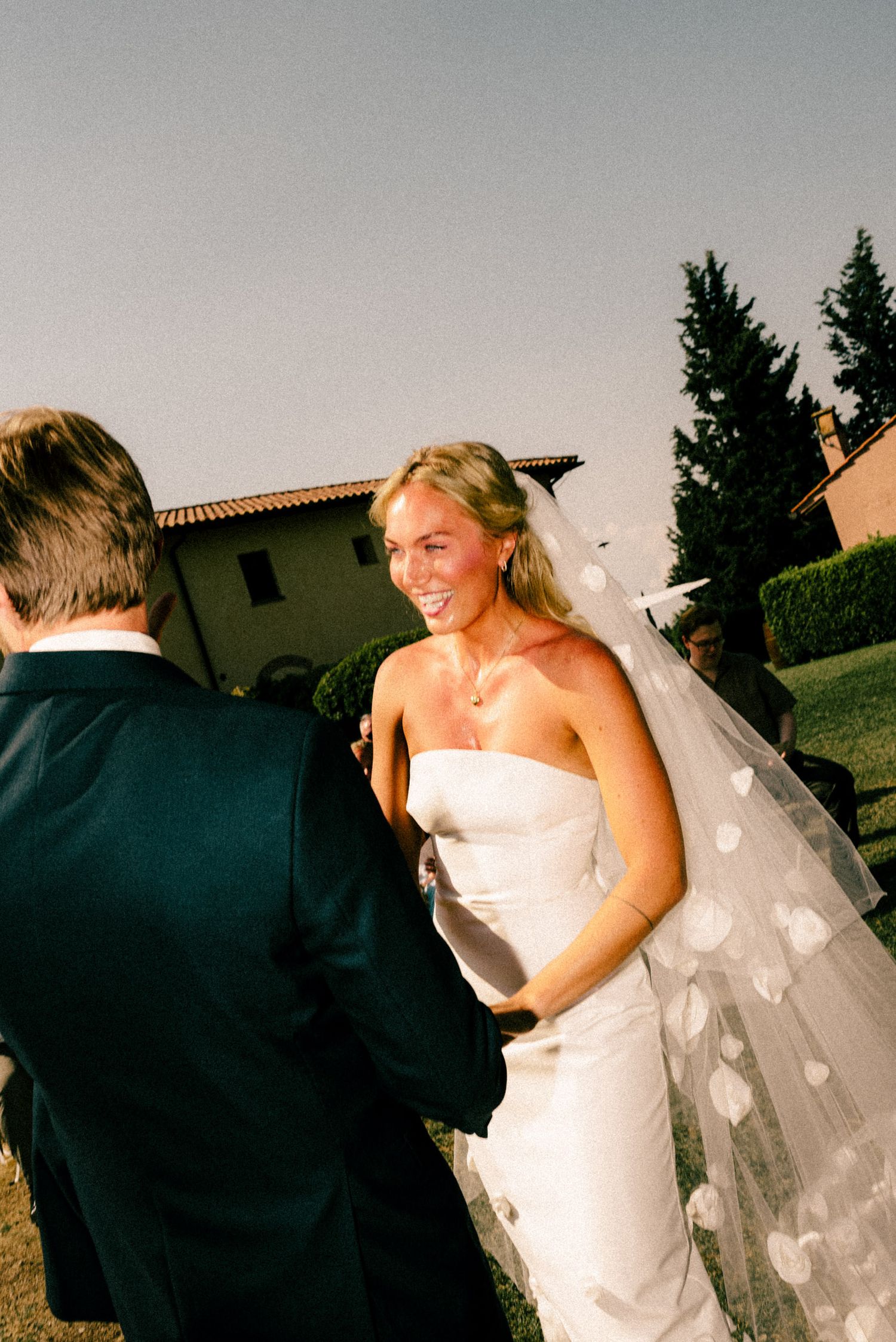 Smiling bride in strapless white gown with heart-print veil stands with groom outdoors at sunny wedding ceremony.