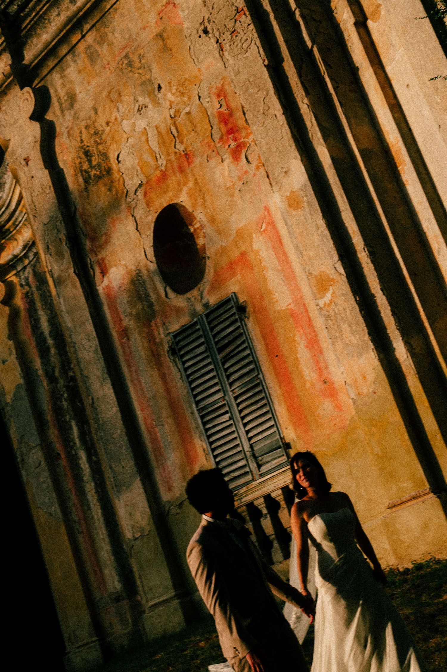 Couple sharing a romantic moment against a weathered Italian building wall with shuttered window in warm golden light.