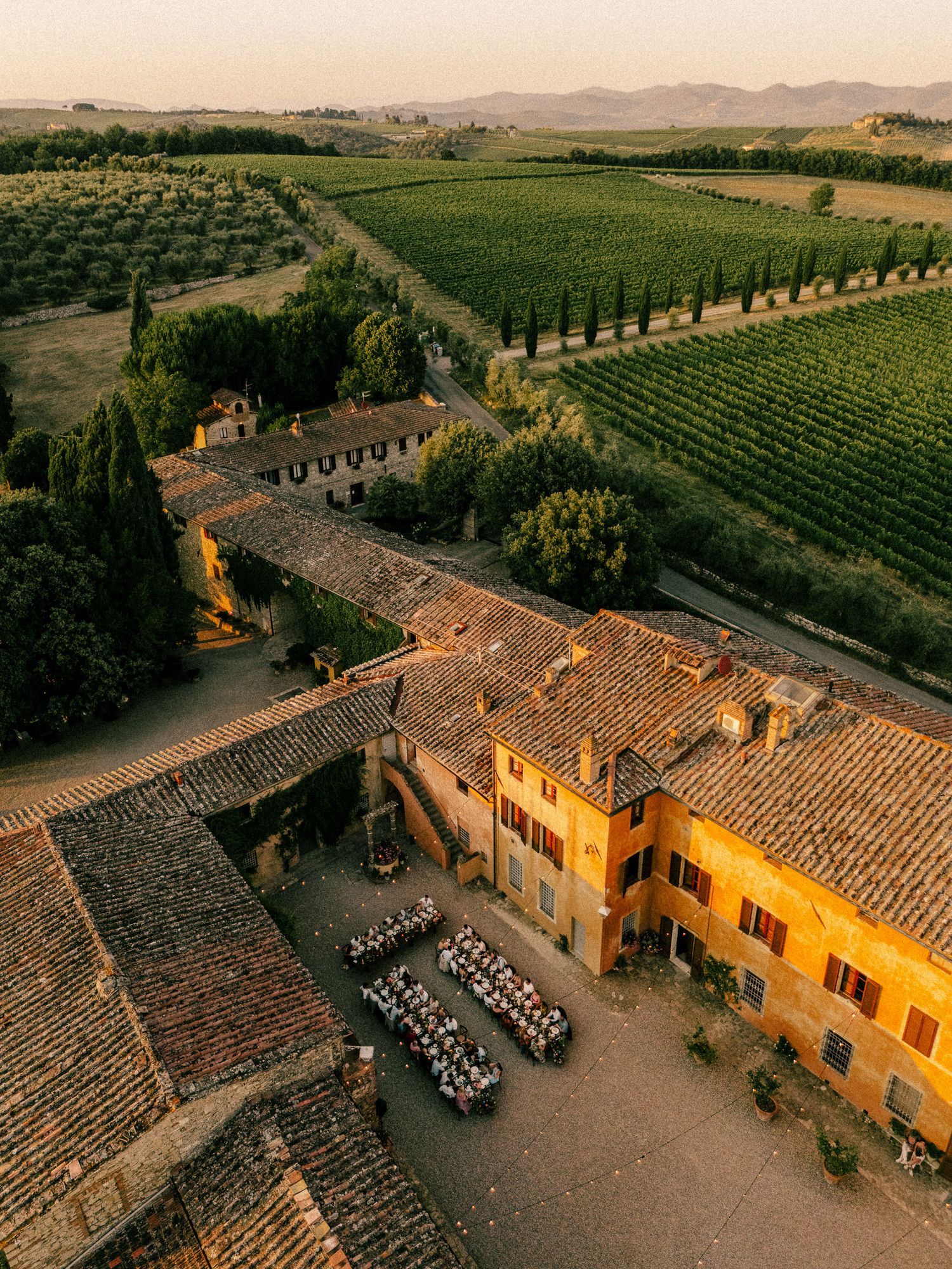 Aerial view of a Tuscan villa with terracotta roofs, outdoor dining, vineyard, and cypress trees at golden hour.