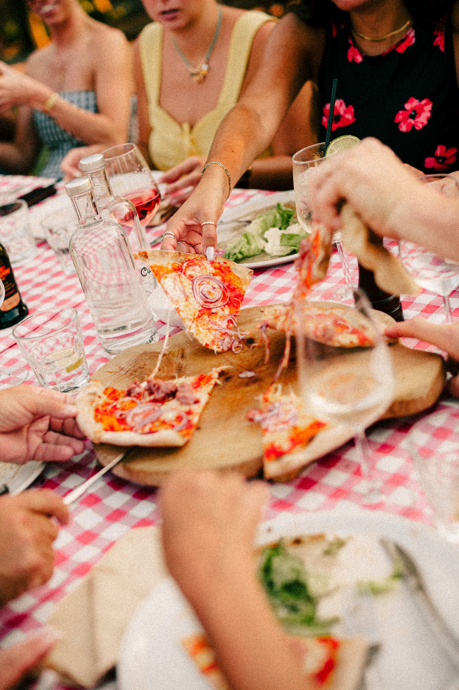 Friends sharing slices of pizza on a wooden board at a red checkered tablecloth table with water glasses and salad.