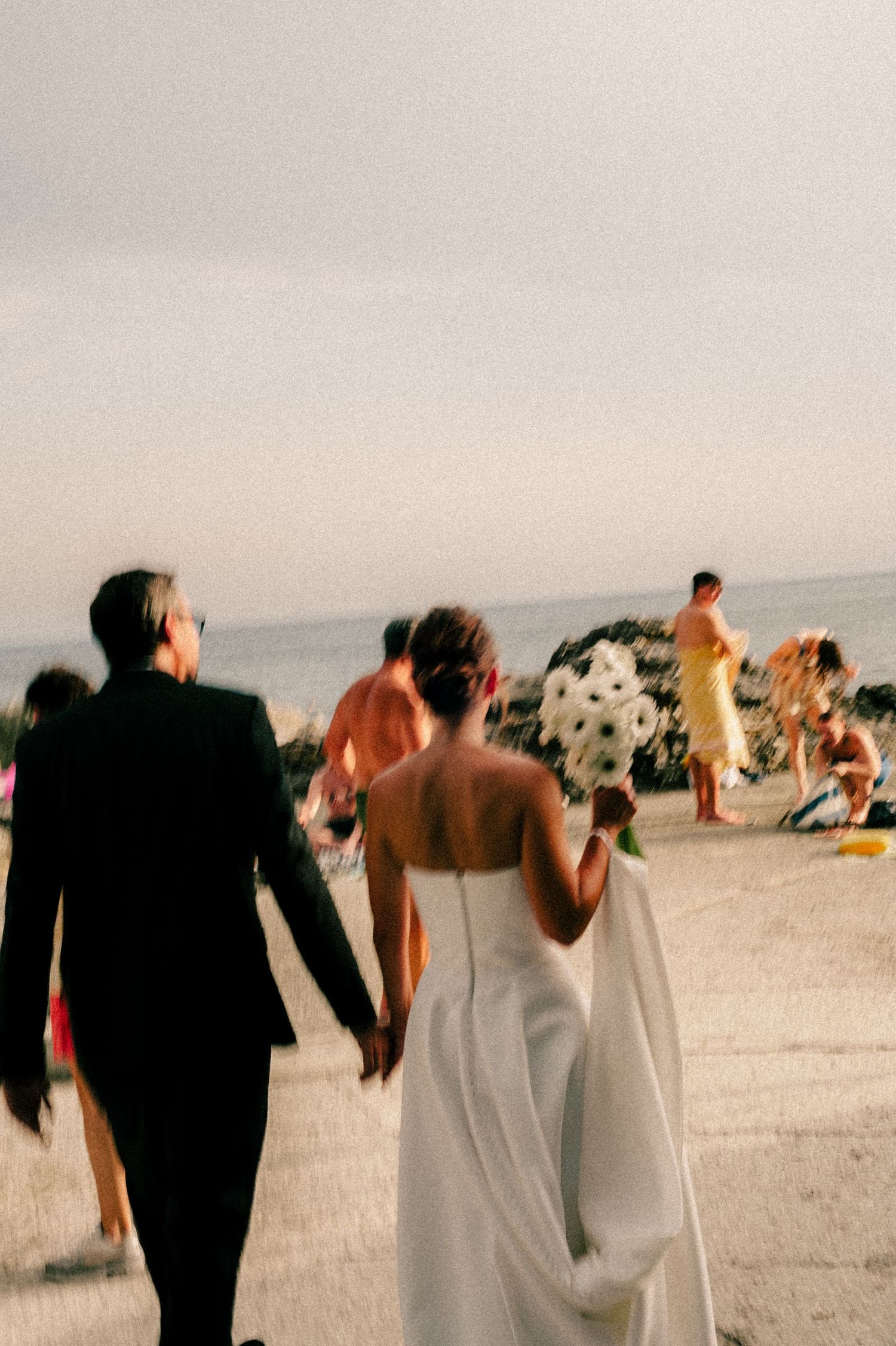 Bride and groom walk hand-in-hand on a beach, bride holding black and white bouquet, guests in background.