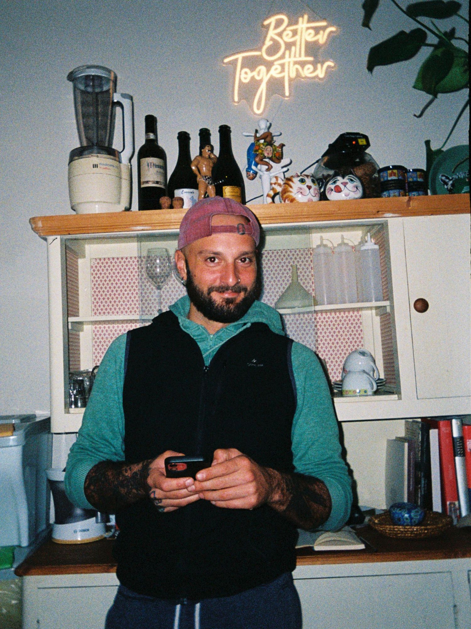 Bearded man in teal sweater and pink cap holds phone, standing before a cabinet with a neon 'Better Together' sign.