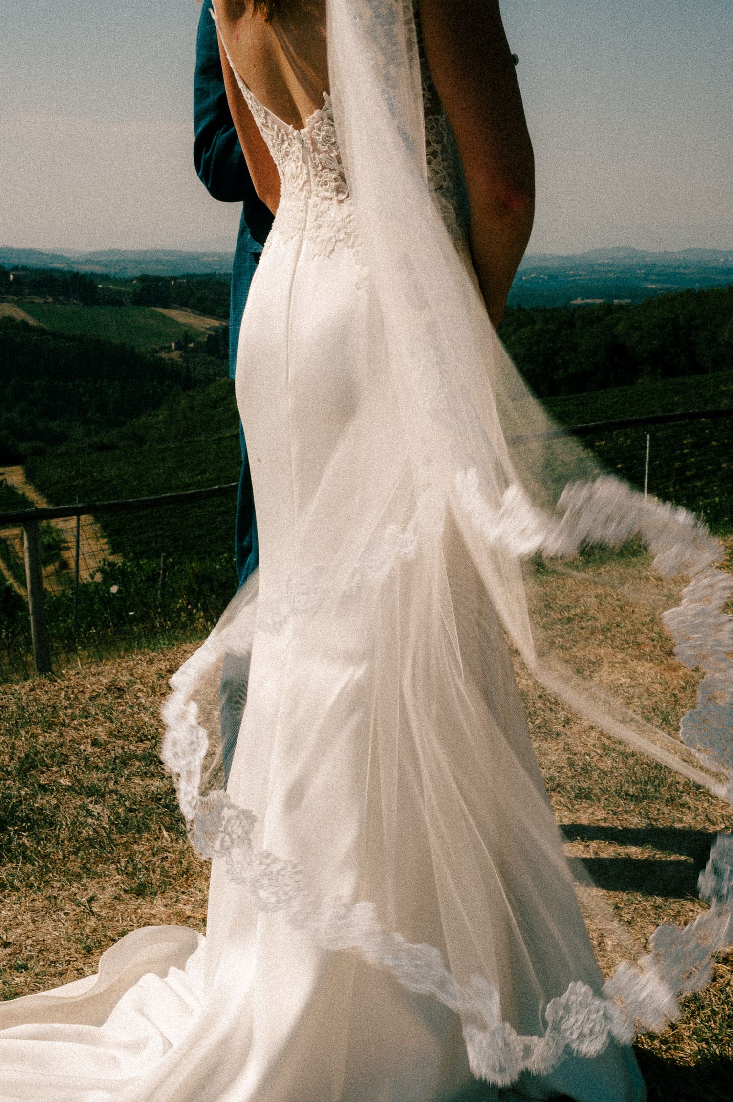Bride in elegant backless lace wedding gown with flowing veil, standing with groom overlooking scenic Tuscan hills.