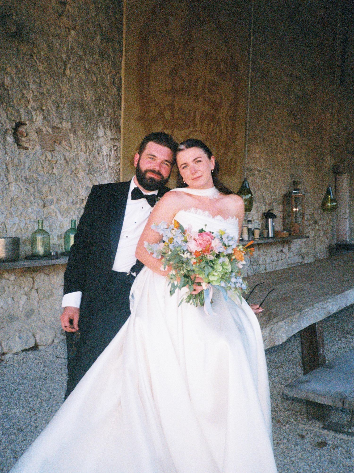 A newlywed couple shares a romantic moment together in front of a rustic stone wall during their wedding celebration.