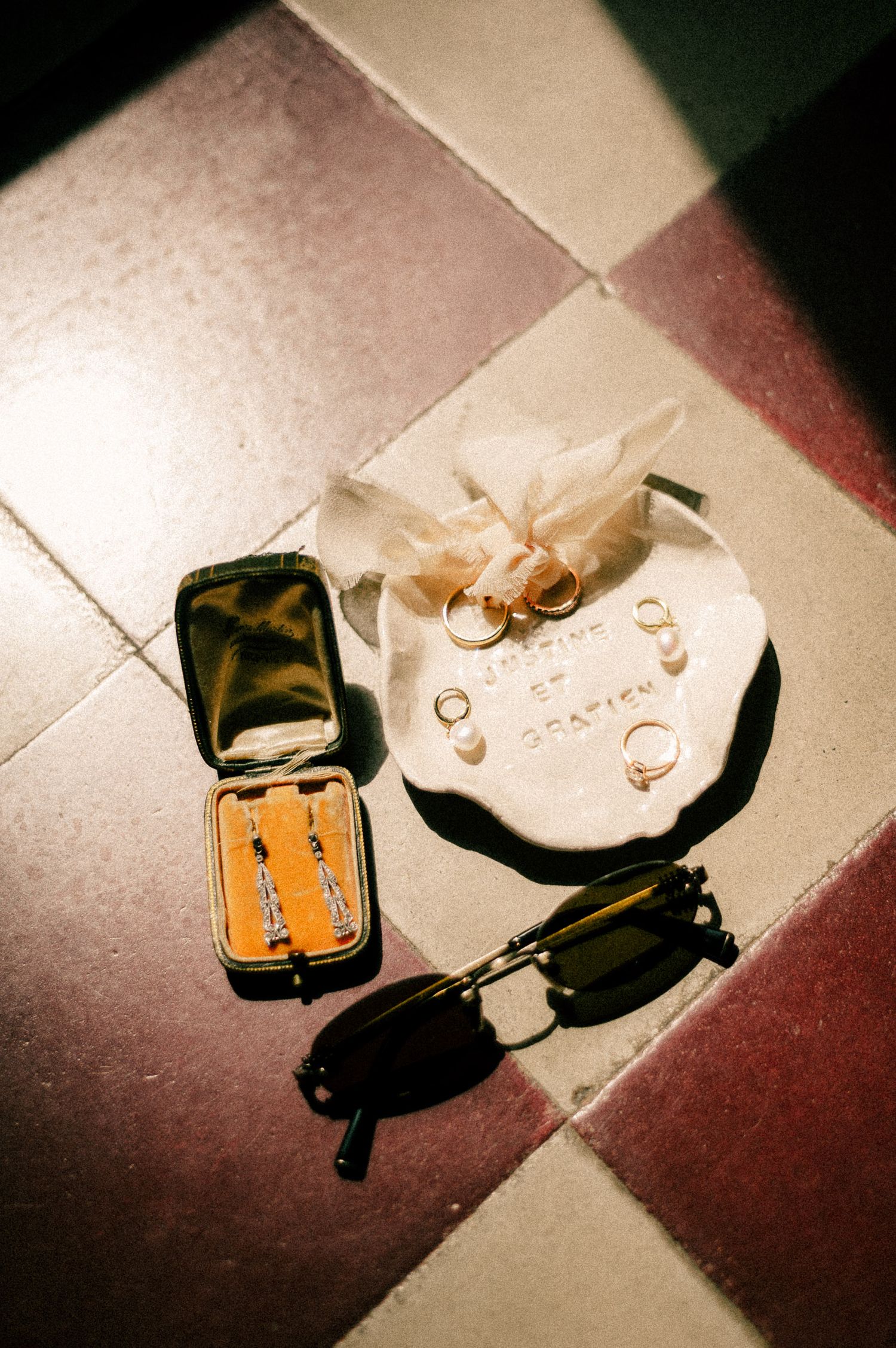 Wedding rings on ceramic dish with pearl accents, vintage jewelry box, and retro sunglasses arranged on colorful tile floor.