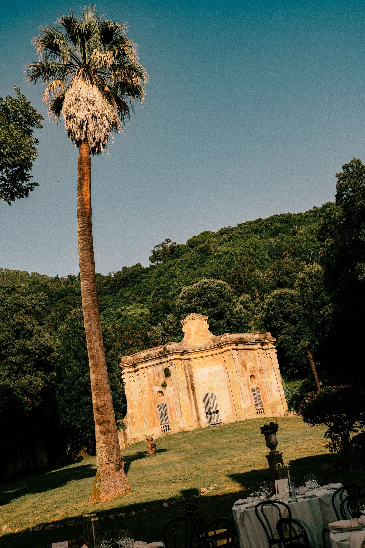 Historic baroque chapel beside a tall palm tree on lush green grounds with forested hills at golden hour.