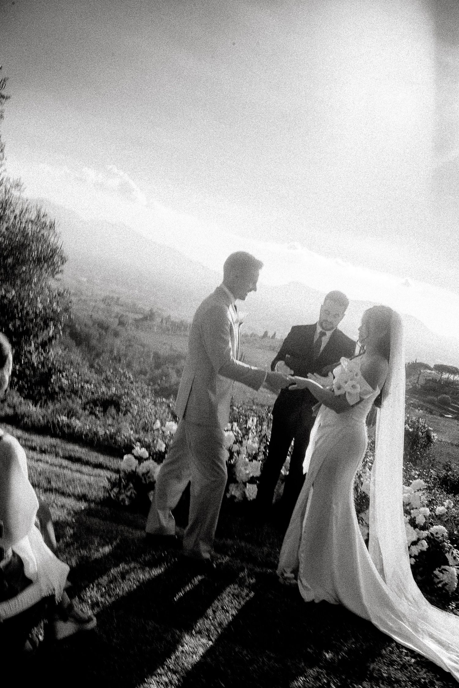 A black and white wedding ceremony takes place on a hillside with mountains visible in the misty background.
