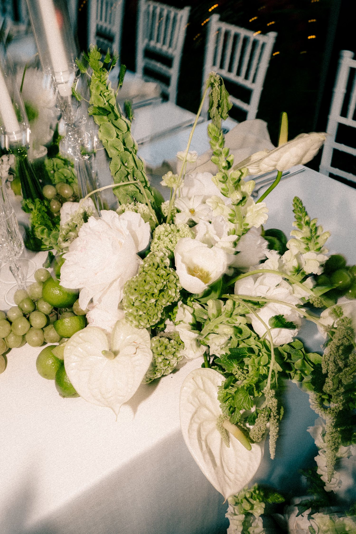 Elegant white floral centerpiece with green accents including anthurium and grapes on white tablecloth.