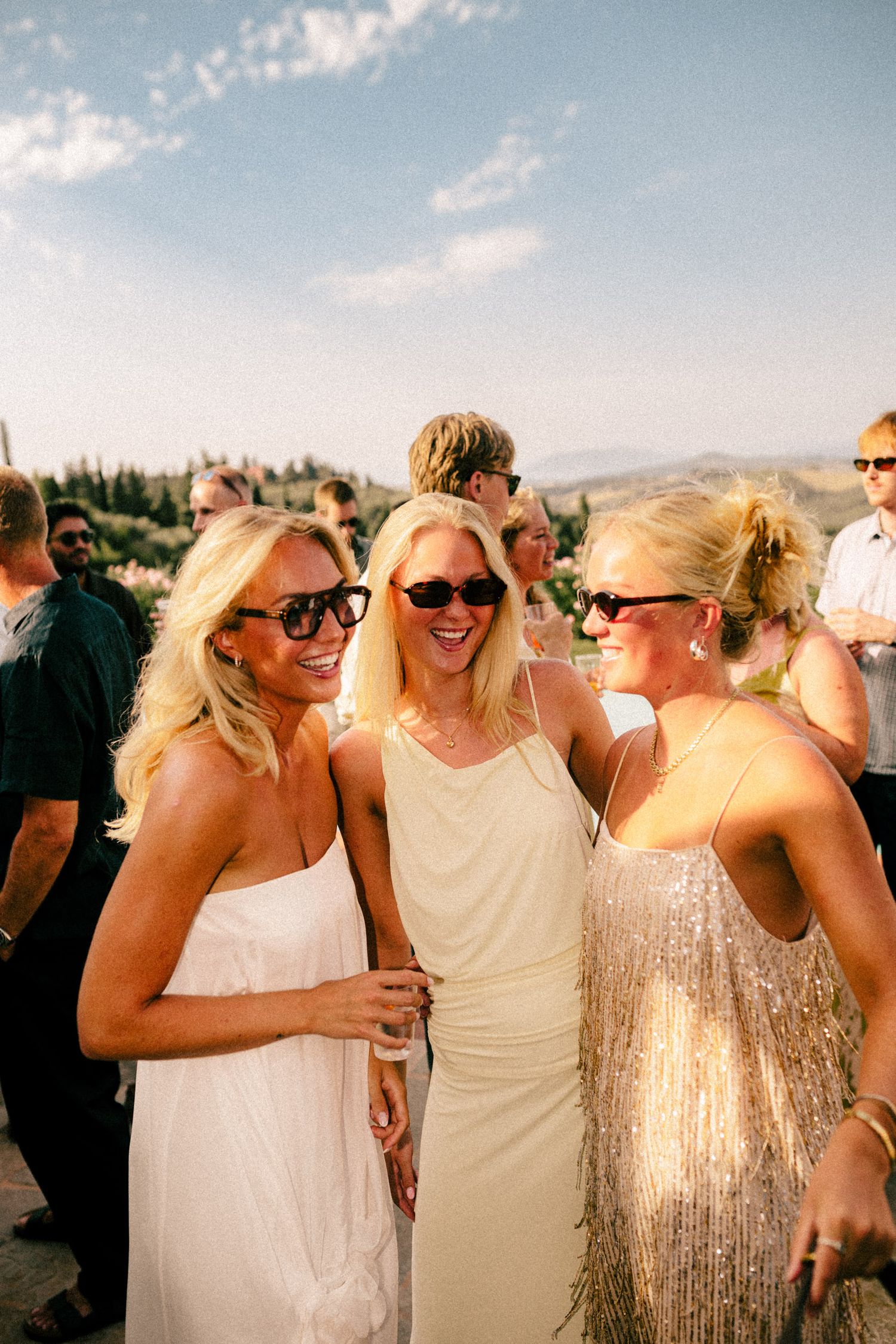 Three blonde women in elegant dresses and sunglasses laughing together at an outdoor wedding reception.