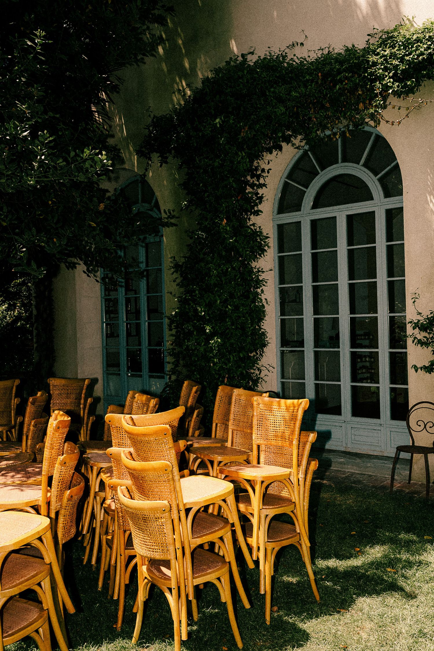 Wooden rattan chairs stacked beside outdoor dining tables on a lawn, beside a white building with arched windows.