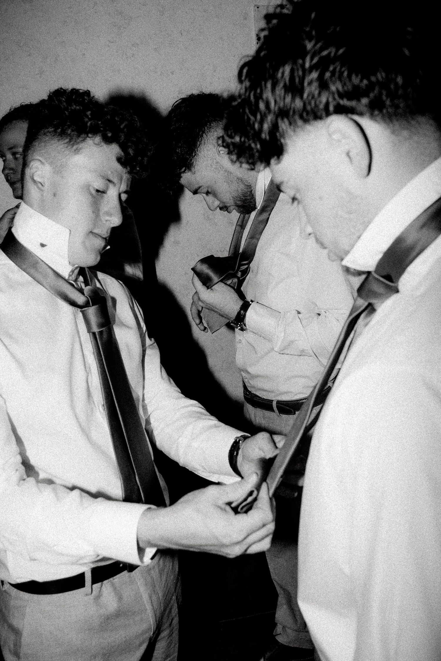 Black and white photo of groomsmen helping each other with ties and suspenders before a wedding ceremony.