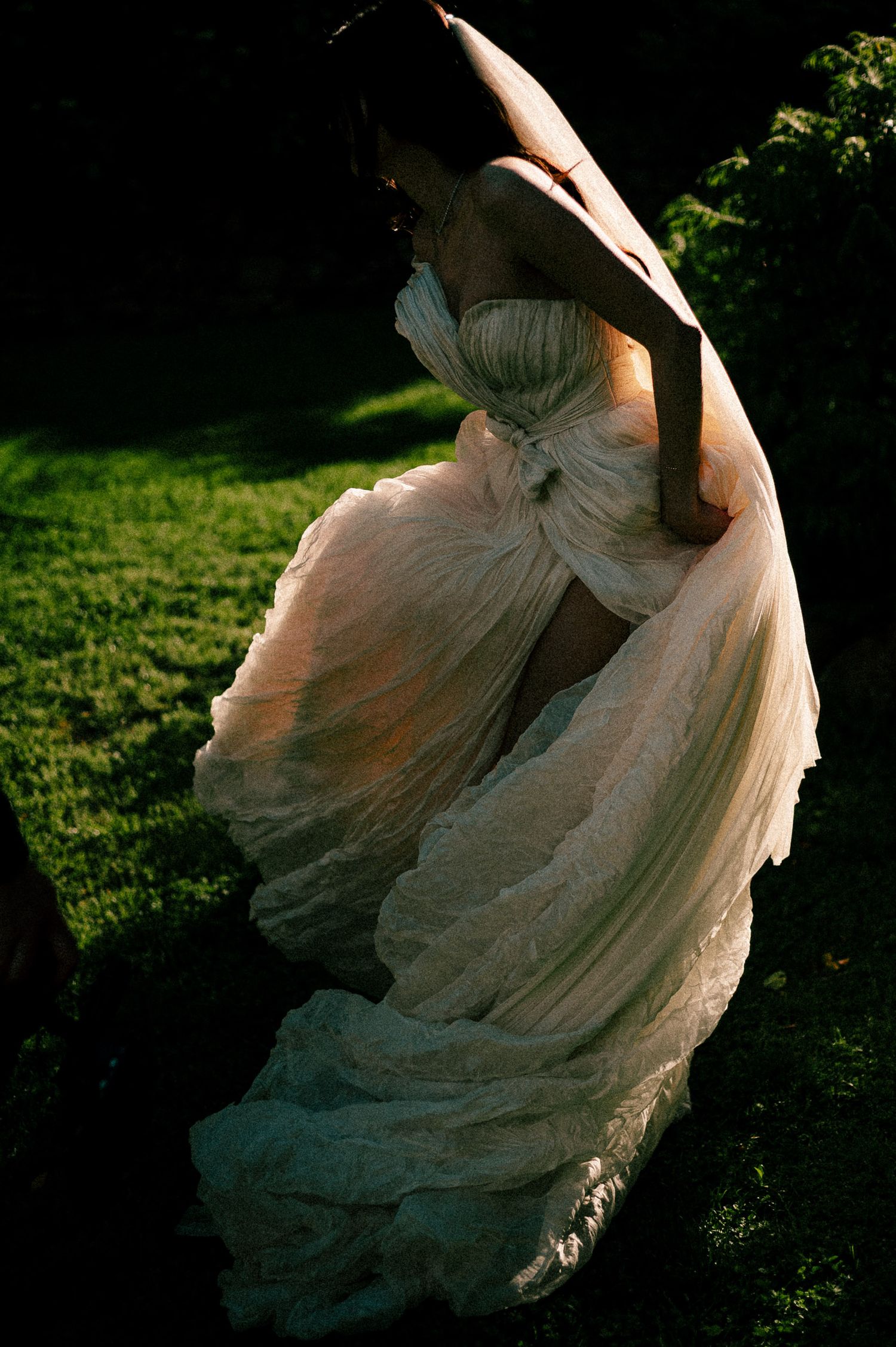 A bride twirls in a flowing strapless white gown on a sunlit green lawn, captured in dramatic light and shadow.