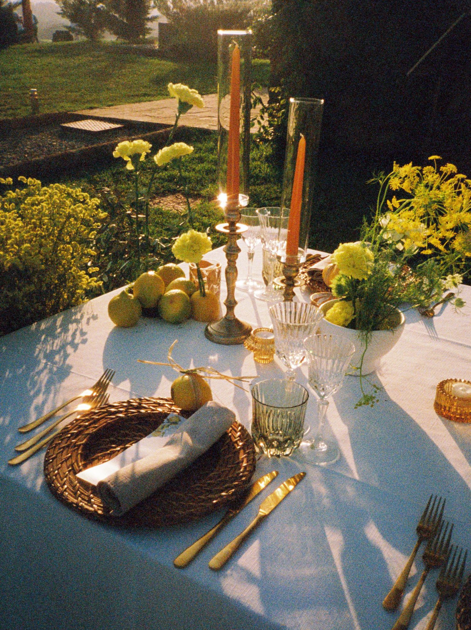 Elegant place settings with woven chargers, gold flatware, and yellow floral centerpieces on a white tablecloth in natural light.